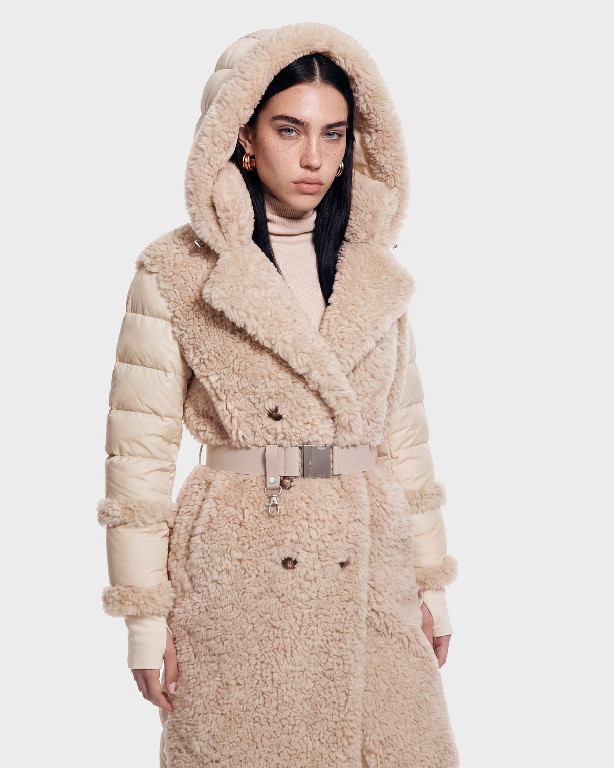 A woman wearing the NOELLE - 8126971 Cream plush faux fur coat by Rudsak, featuring a hooded, belted design and quilted sleeves, stands against a plain light background.
