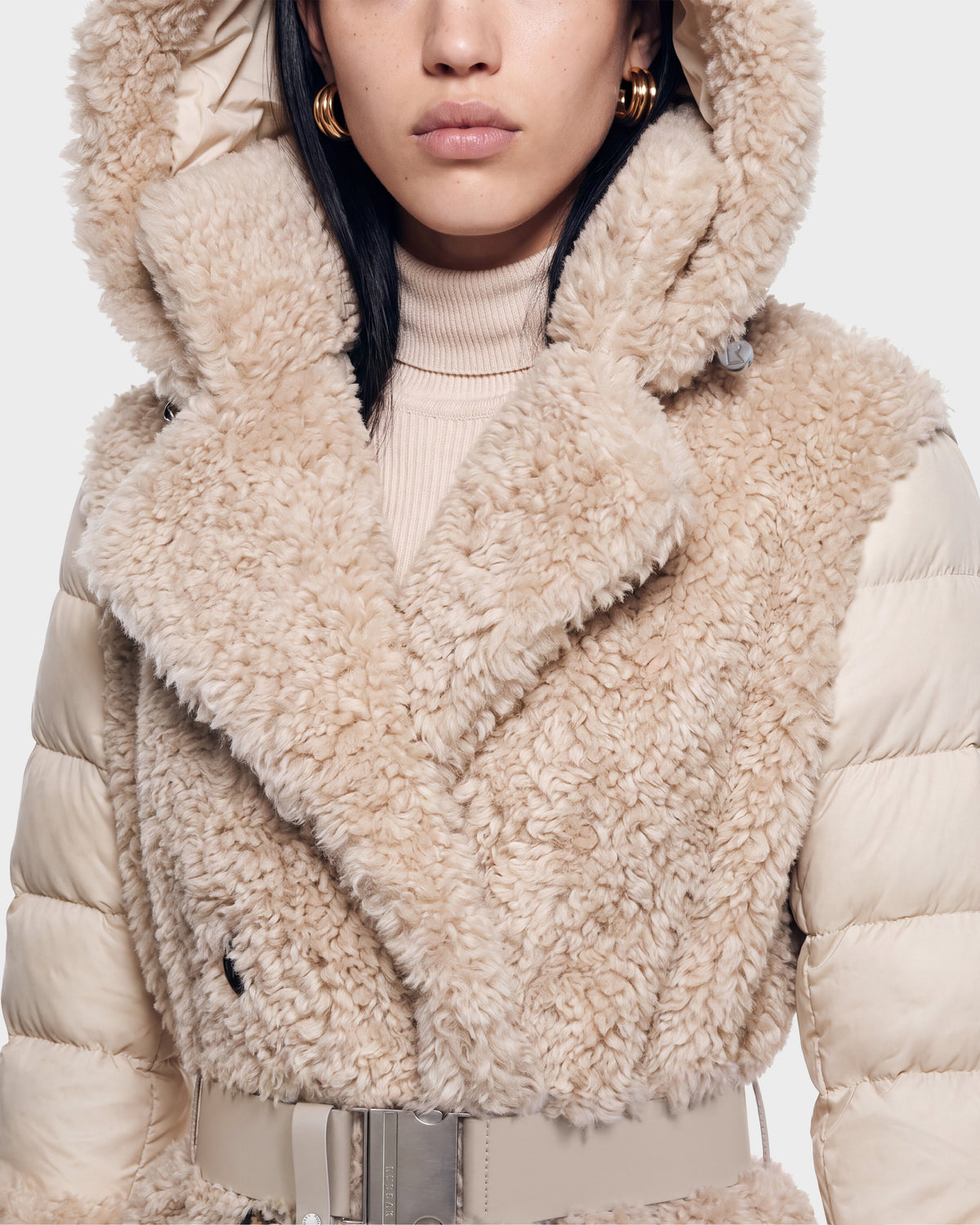 A woman wearing the Rudsak NOELLE - 8126971 Cream faux fur coat with a large hood, puffy sleeves, wide collar, turtleneck sweater, hoop earrings, and a wide belt. The photo is cropped to show her mid-face and upper torso.