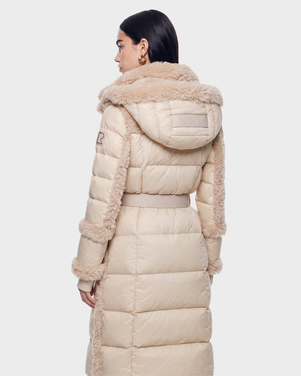 A woman with dark hair models the Rudsak NOELLE - 8126971 Cream, a long down coat featuring a fur-trimmed hood and sleeves, as she faces away from the camera against a plain, light background.