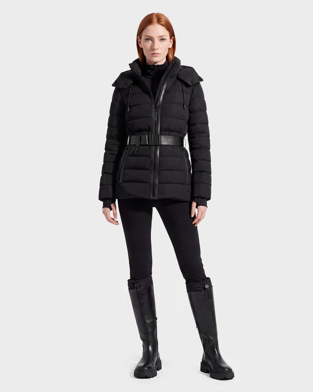 A woman with straight red hair wears the Rudsak PELIN S - 8124813 Black belted quilted puffer jacket, paired with black pants and knee-high black boots, standing against a plain light gray background.