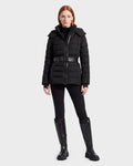 A woman with straight red hair wears the Rudsak PELIN S - 8124813 Black belted quilted puffer jacket, paired with black pants and knee-high black boots, standing against a plain light gray background.