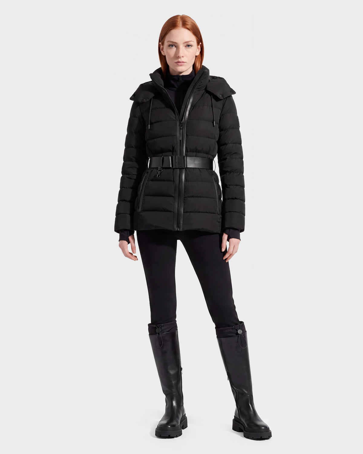 A woman with straight red hair wears the Rudsak PELIN S - 8124813 Black belted quilted puffer jacket, paired with black pants and knee-high black boots, standing against a plain light gray background.