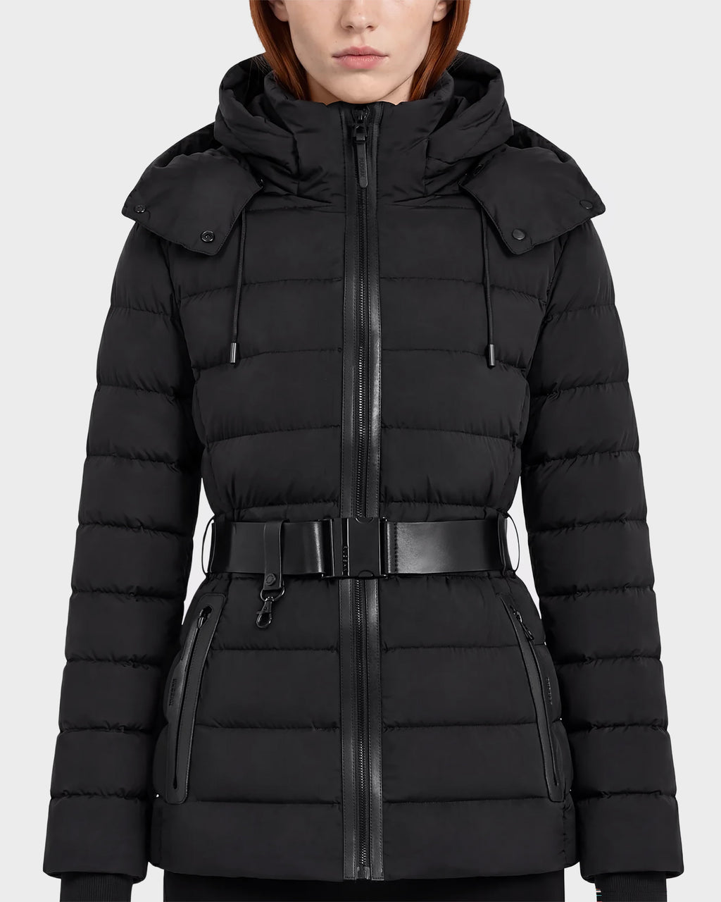 A woman wearing the Rudsak PELIN S - 8124813 Black coat, a belted quilted puffer jacket with a hood, zippered front, and zippered pockets, stands against a plain light background.