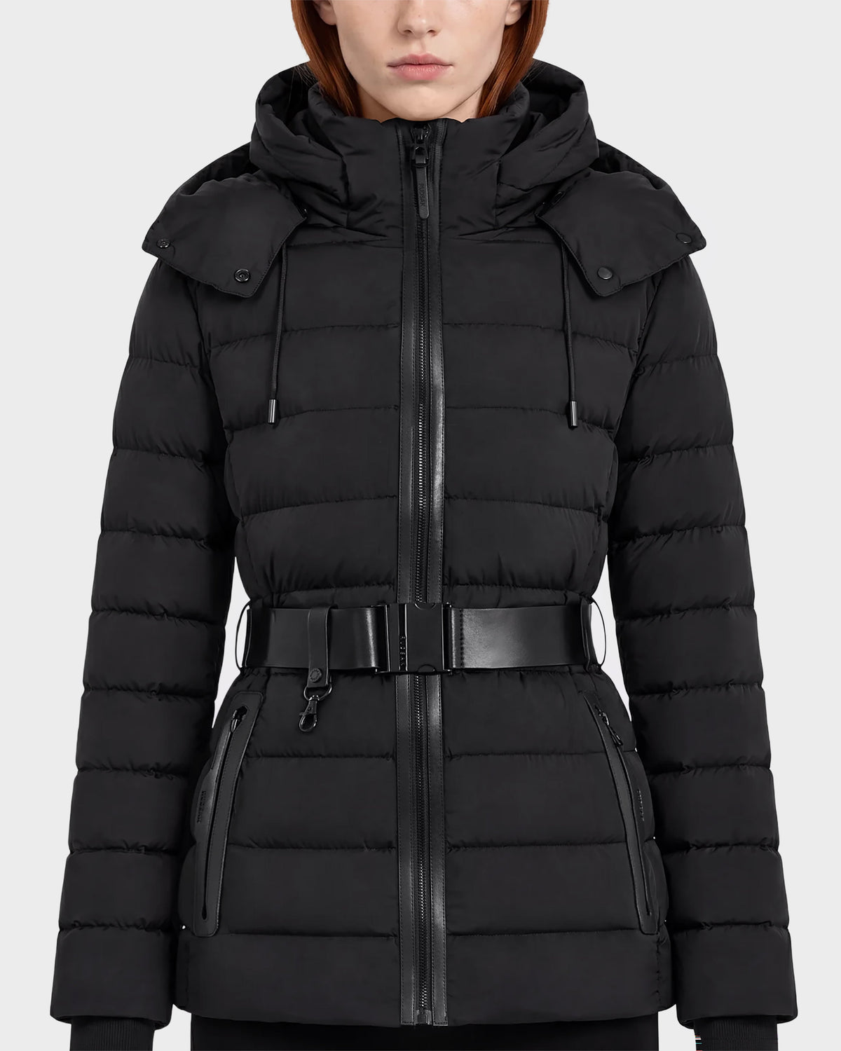 A woman wearing the Rudsak PELIN S - 8124813 Black coat, a belted quilted puffer jacket with a hood, zippered front, and zippered pockets, stands against a plain light background.
