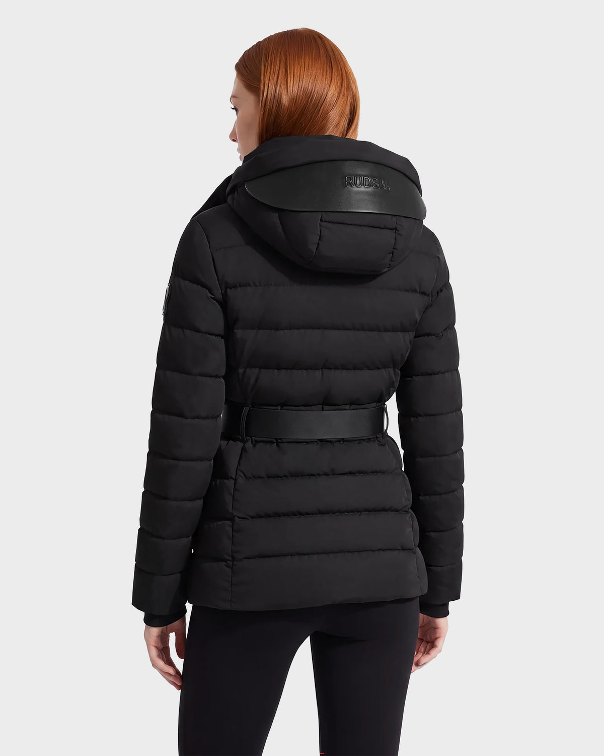 A person with long red hair is seen from the back against a plain light background, wearing the Rudsak PELIN S - 8124813 Black belted, quilted puffer jacket with a hood.