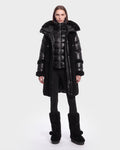 A person stands facing forward, dressed in the Rudsak SHELLEY - 8126729 Black long coat layered over a shiny black puffer jacket, paired with black leggings and tall black winter boots with pom-poms. The background is plain and light-colored.