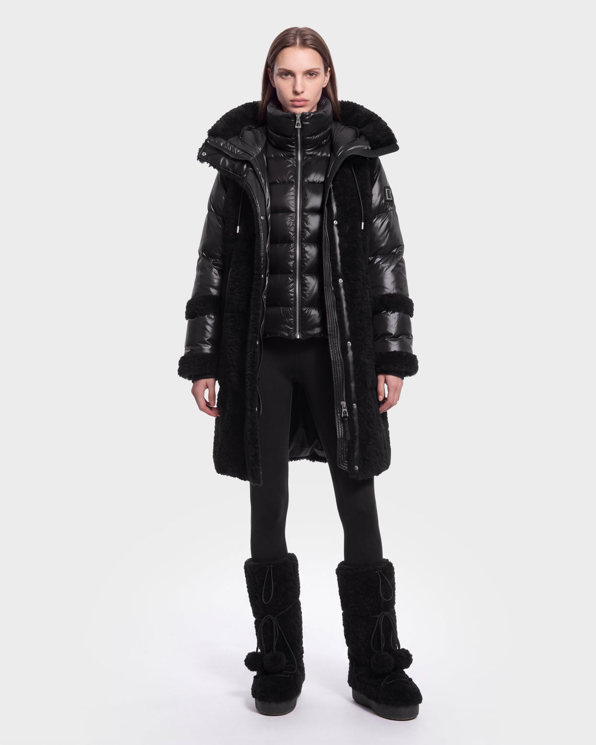 A person stands facing forward, dressed in the Rudsak SHELLEY - 8126729 Black long coat layered over a shiny black puffer jacket, paired with black leggings and tall black winter boots with pom-poms. The background is plain and light-colored.