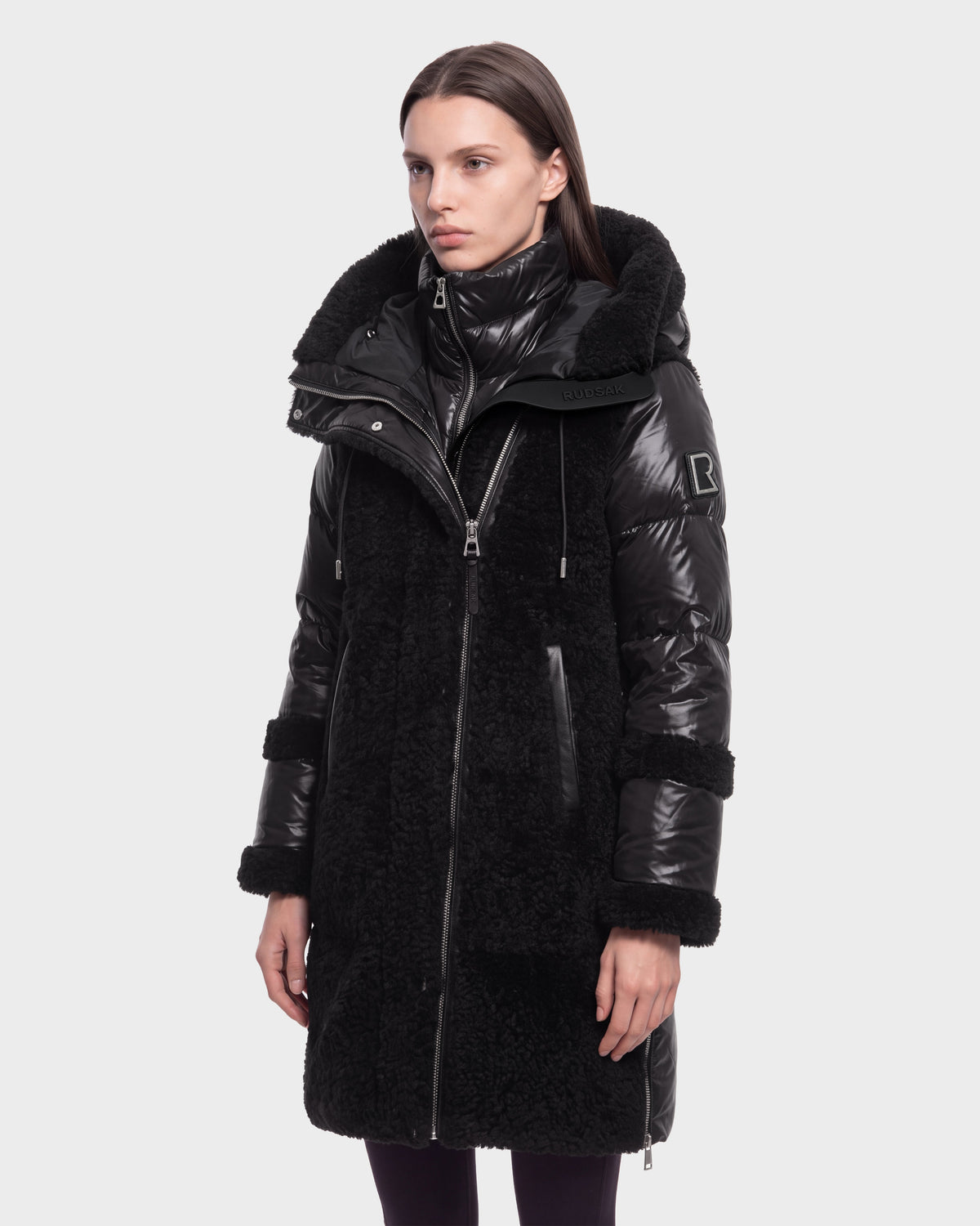 A woman stands facing forward with a neutral expression against a plain white background, wearing the Rudsak SHELLEY - 8126729 Black winter coat, which is long, black, shiny, and textured with a high collar and hood.