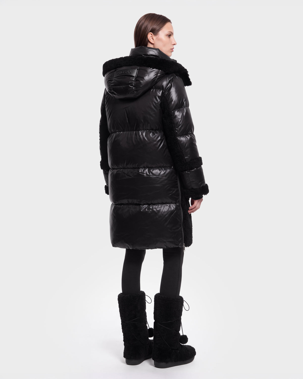 A woman with straight brown hair stands facing away against a plain light background, dressed in the Rudsak SHELLEY - 8126729 Black long, shiny puffer coat with a hood, paired with black tights and matching black boots.