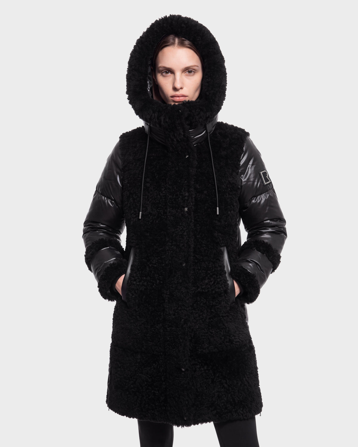 A person wearing the Rudsak SHELLEY - 8126729 Black coat, featuring shiny sleeves and a hood, stands with hands in pockets against a plain light background.