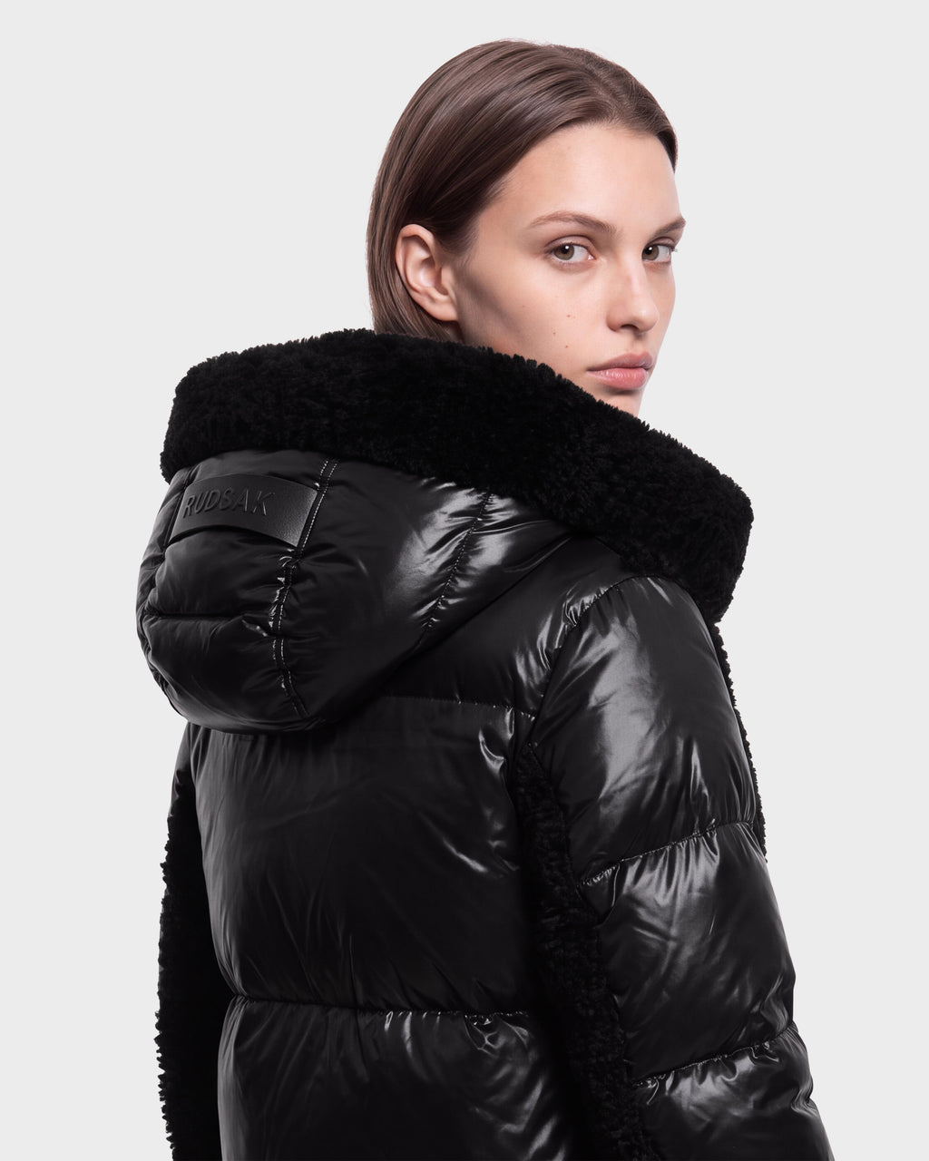 A person with straight brown hair models the Rudsak SHELLEY - 8126729 Black puffer jacket, featuring a shiny finish and faux fur hood, as they look over their shoulder against a plain light background.