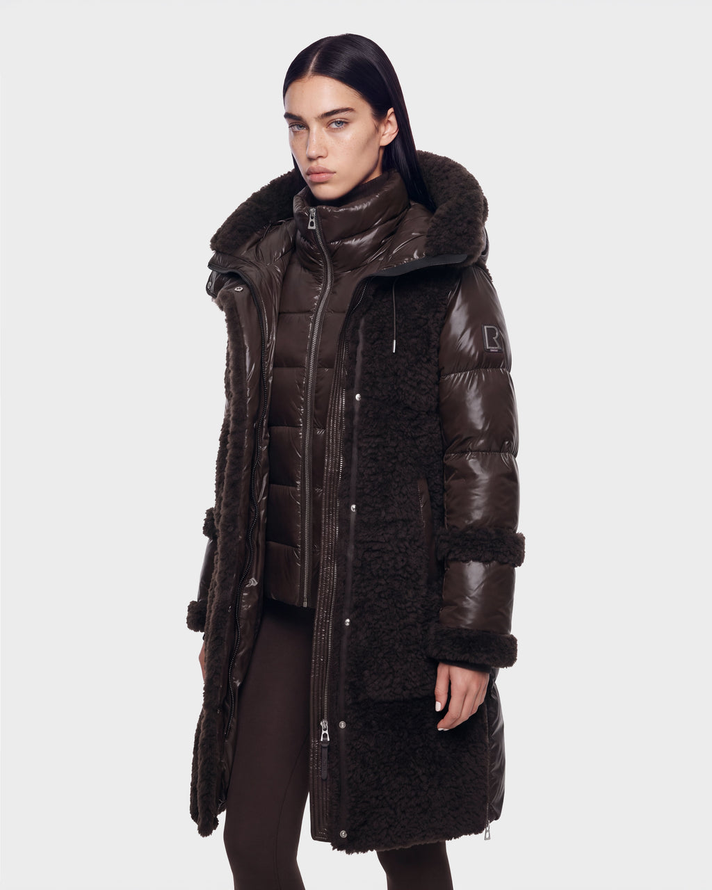 A woman wearing the Rudsak SHELLEY - 8126729 Espresso, a long espresso-colored winter coat featuring a puffy, sherpa-textured design and down insulation, stands against a plain white background, looking ahead with a neutral expression.