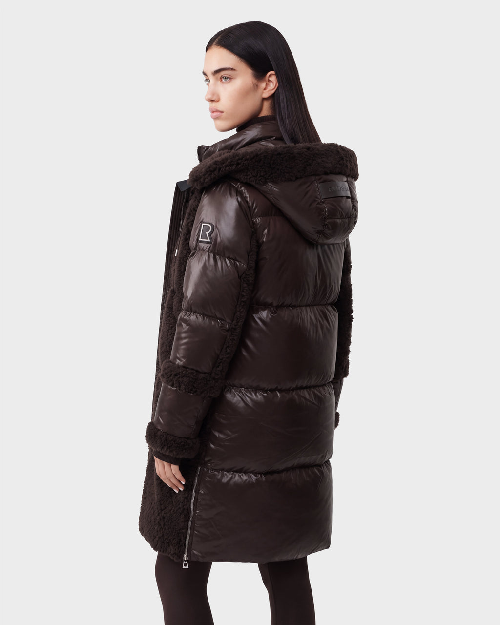 A woman with straight dark hair models the Rudsak SHELLEY - 8126729 Espresso, a long, espresso-colored puffer coat with down insulation and a fur-trimmed hood, standing against a plain light background and facing away at a slight angle.