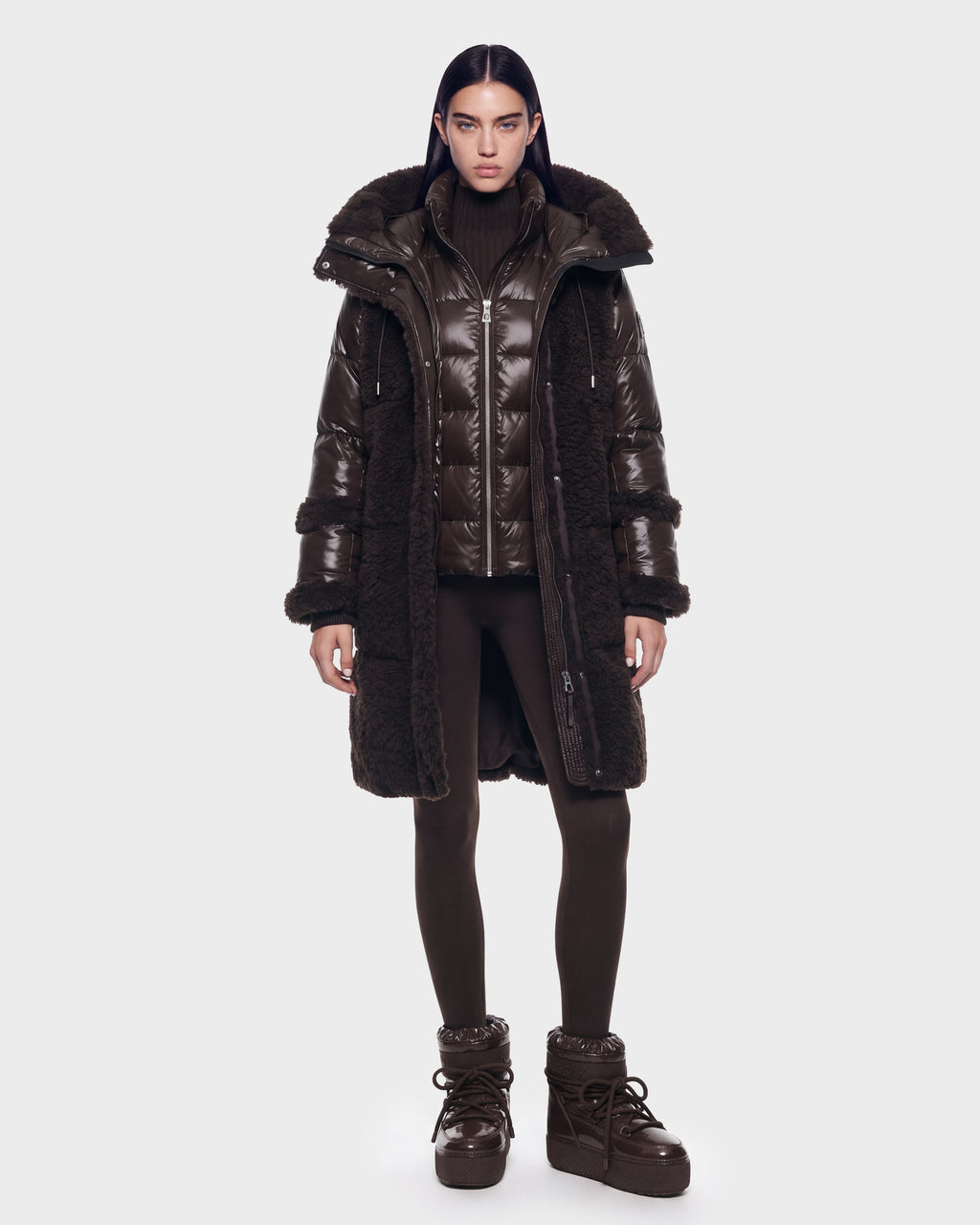 A person stands facing forward against a plain light background, wearing the Rudsak SHELLEY - 8126729 Espresso—a long, dark brown puffer coat with down insulation and fur trim—paired with a matching turtleneck, leggings, and high-top boots for a stylish winter outerwear look.