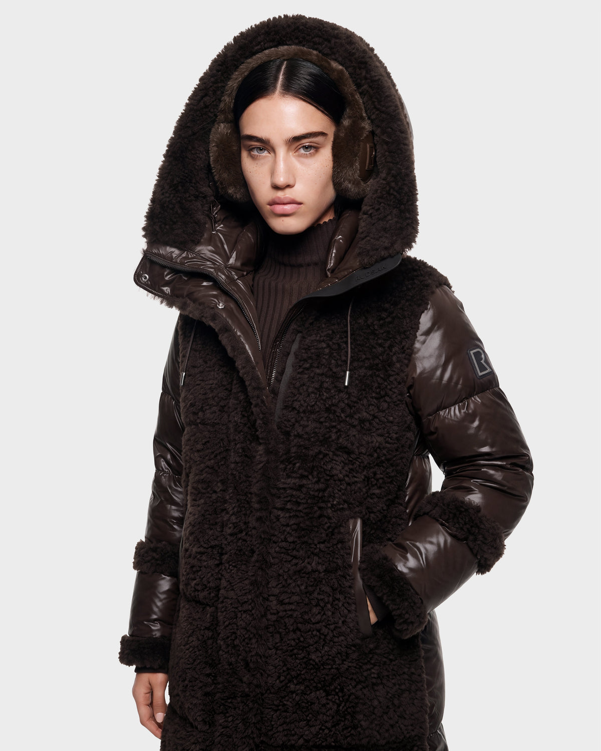 A woman wearing the Rudsak SHELLEY - 8126729 Espresso dark brown hooded winter coat with sherpa-textured panels and ear muffs stands against a plain light background, looking directly at the camera with a neutral expression.