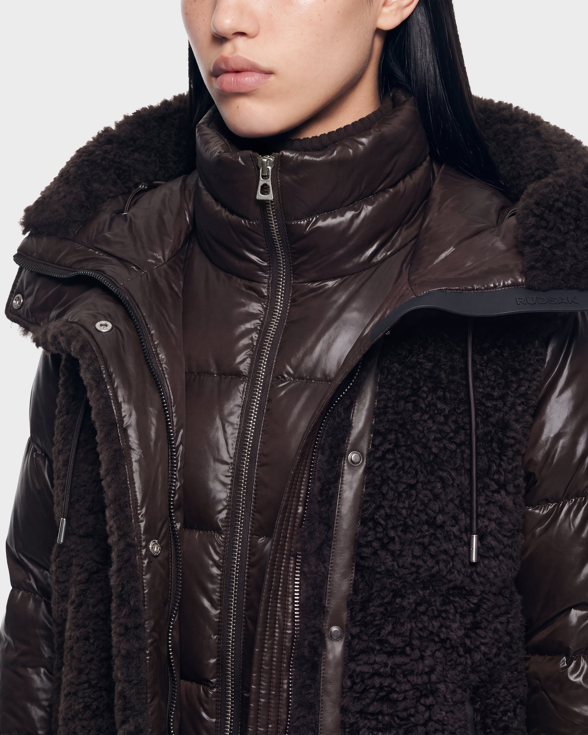 Close-up of a person wearing the Rudsak SHELLEY - 8126729 Espresso puffer jacket, featuring a high collar and a textured, shiny dark brown outer layer. Designed for winter outerwear, this jacket showcases visible zippers and snap buttons. The person’s face is partially visible.