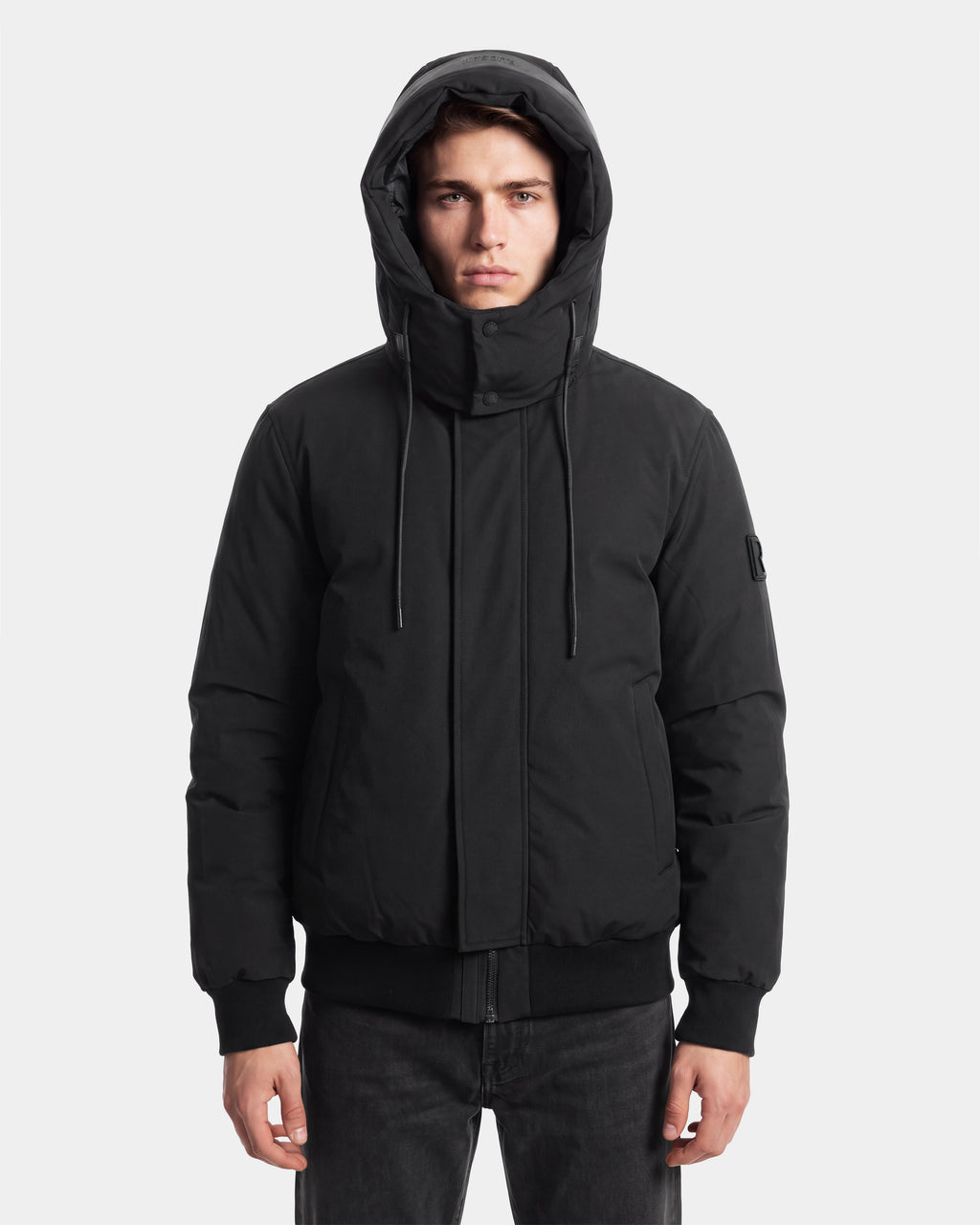 A person wearing the Rudsak WILL - 6126561 Black hooded winter jacket with a high collar stands facing forward with a neutral expression against a plain white background.