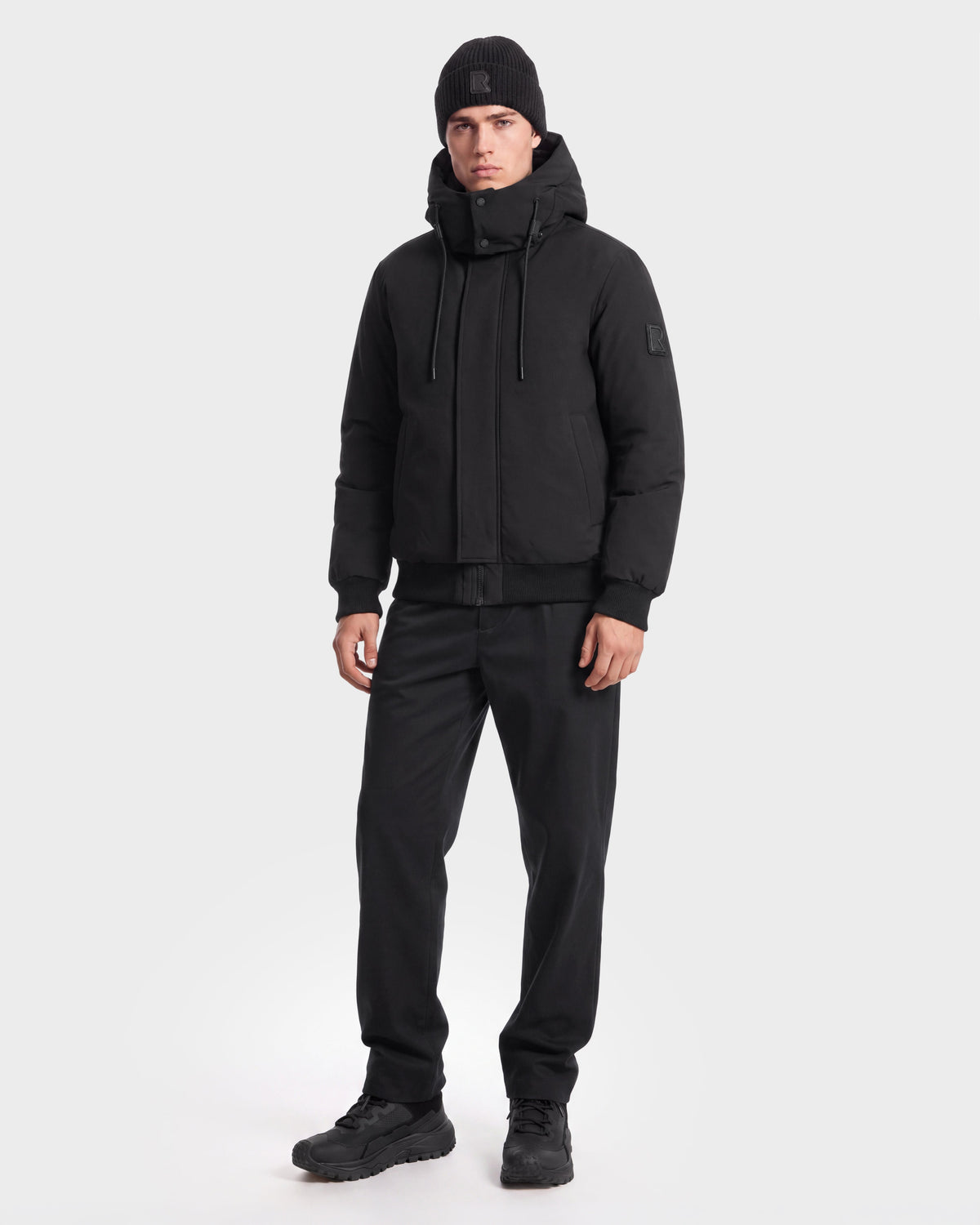 A person wearing the Rudsak WILL - 6126561 Black hooded jacket stands against a plain white background, paired with a black beanie, black pants, and black shoes.