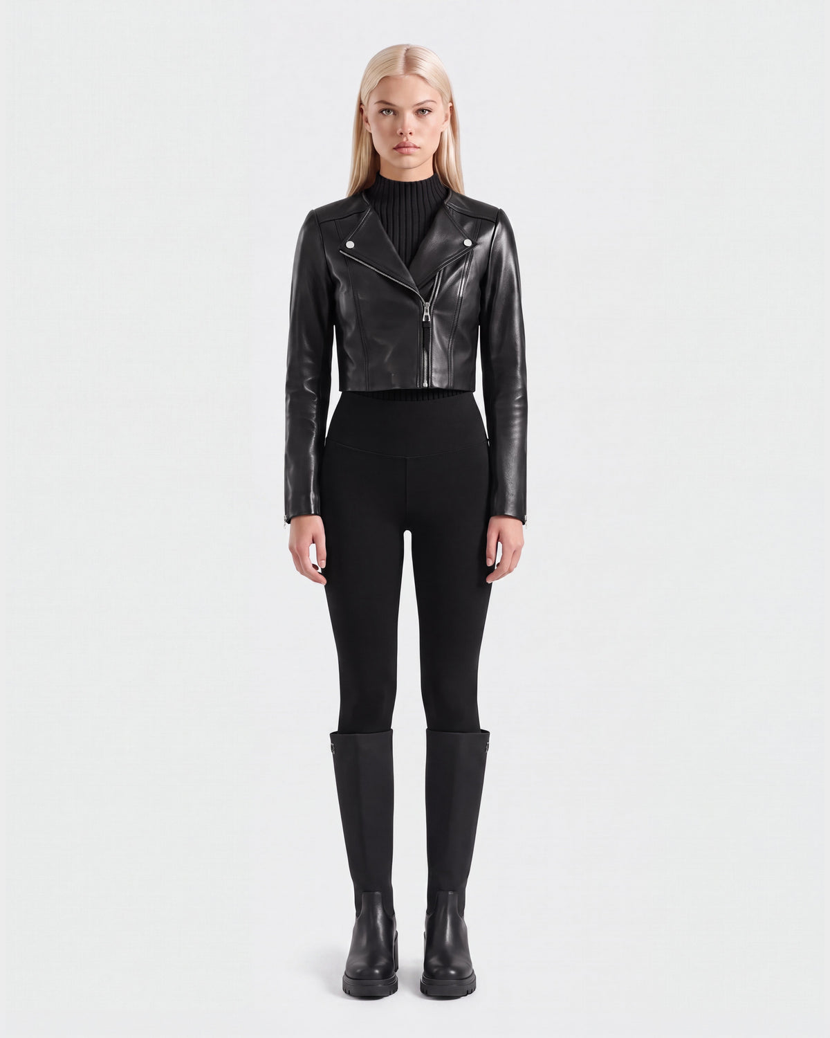A woman stands facing forward against a plain white background, wearing the WINNI - 8124053 Black cropped leather biker jacket by Rudsak, paired with black fitted pants, a black ribbed turtleneck, and black knee-high boots.