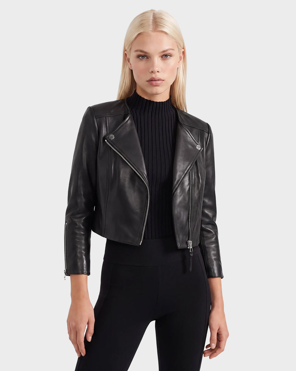 A woman with straight blonde hair wears the Rudsak WINNI - 8124053 Black cropped leather biker jacket over a black ribbed top and black pants, standing against a plain light gray background.