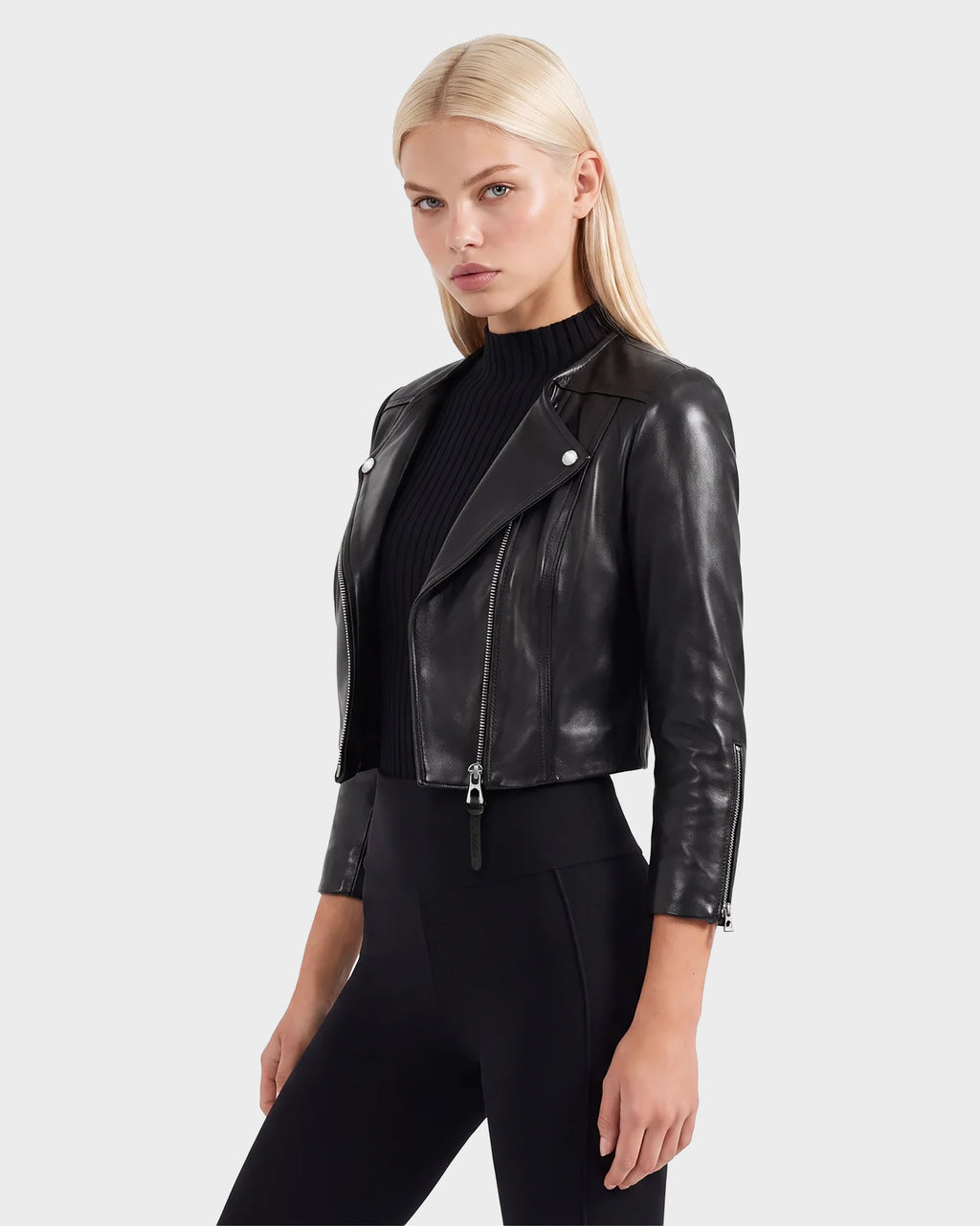 A woman with straight blonde hair wears the WINNI - 8124053 Black fitted women's leather jacket by Rudsak over a black ribbed top and black pants, standing against a plain light gray background.