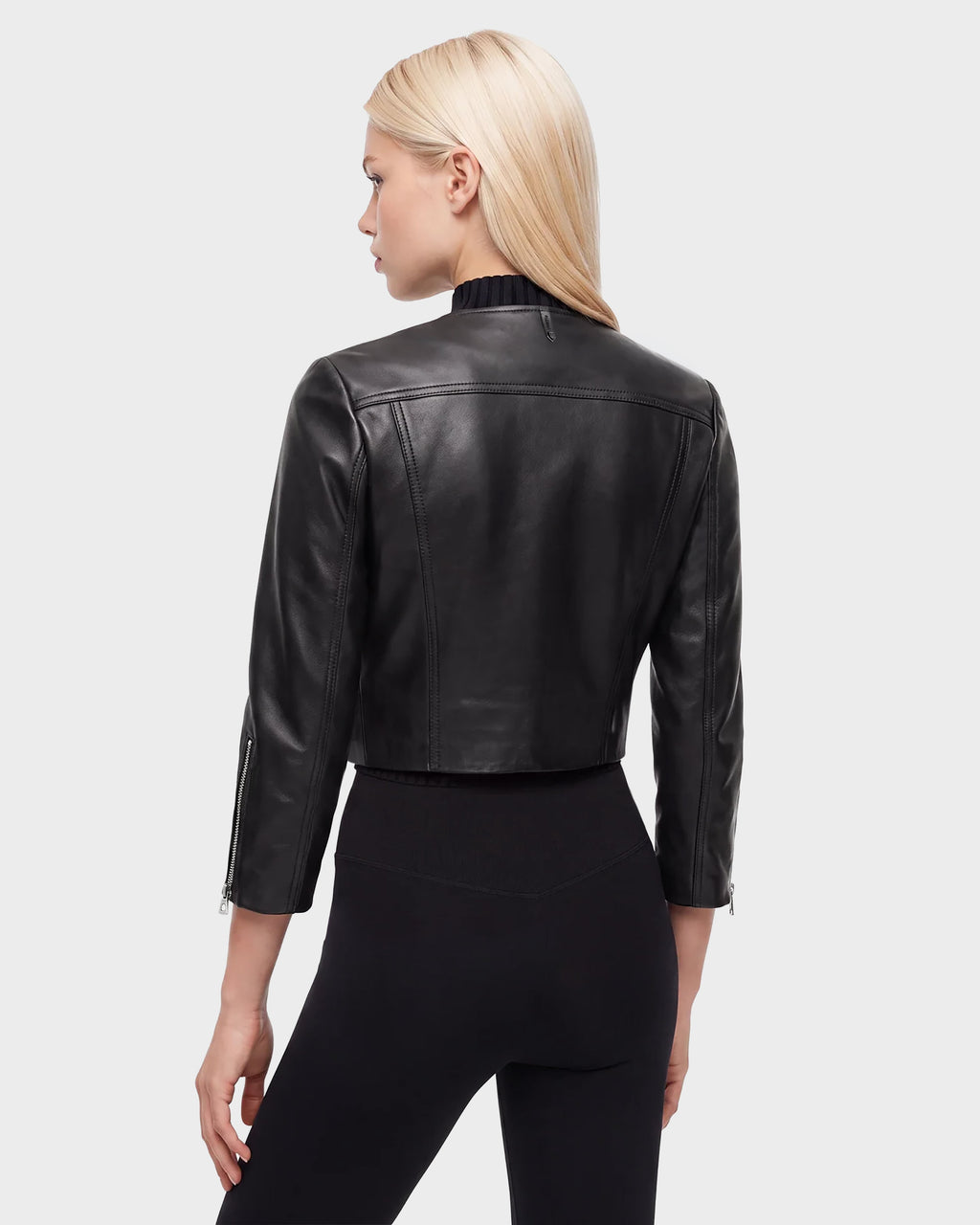 A woman with straight blonde hair faces away, wearing the WINNI - 8124053 Black cropped leather biker jacket by Rudsak and black pants against a plain, light background.