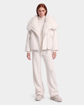 A person stands against a white background wearing Rudsak’s ZARIA - 8126999 Vanilla: an oversized winter puffer with a removable faux-fur collar, paired with a turtleneck sweater, loose pants, and chunky shoes.