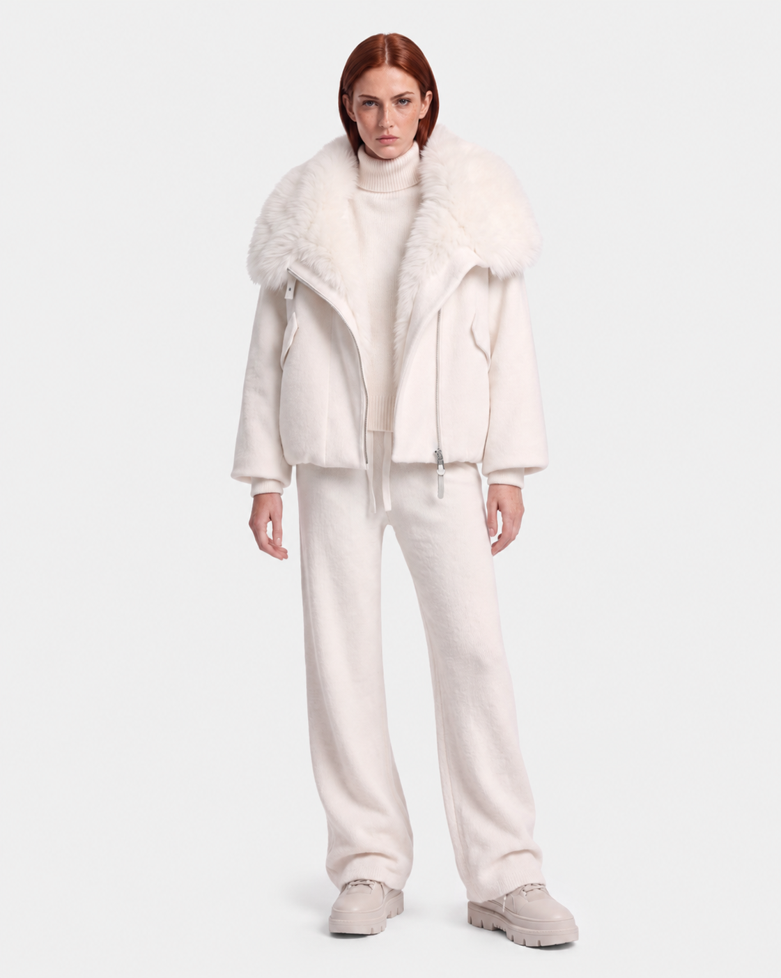 A person stands against a white background wearing Rudsak’s ZARIA - 8126999 Vanilla: an oversized winter puffer with a removable faux-fur collar, paired with a turtleneck sweater, loose pants, and chunky shoes.