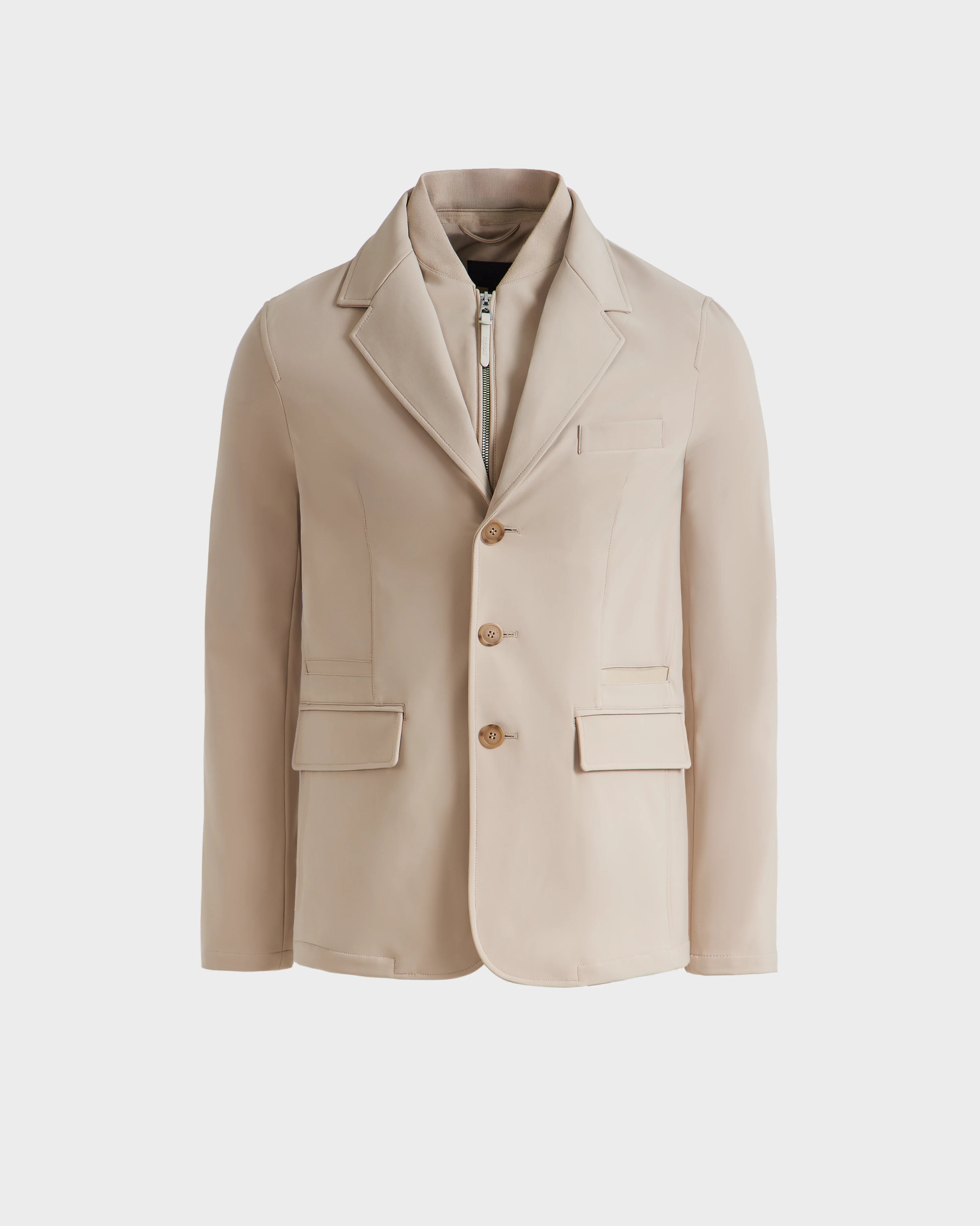 The Rudsak KEITH - 6125045 Almond is a beige blazer-style jacket with notch lapels, three brown buttons, two front flap pockets, a chest pocket, and an inner zip-up layer—modern tailoring shown on a white background.