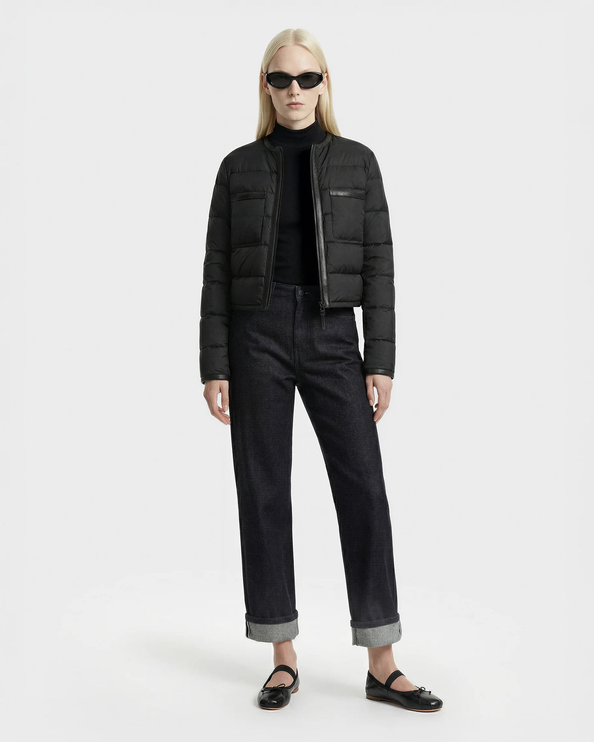 A person with long blonde hair wears black sunglasses and the Rudsak MARKLE - 8125033 Black women’s cropped down jacket, paired with a black turtleneck, dark cuffed jeans, and black flats against a plain white background.