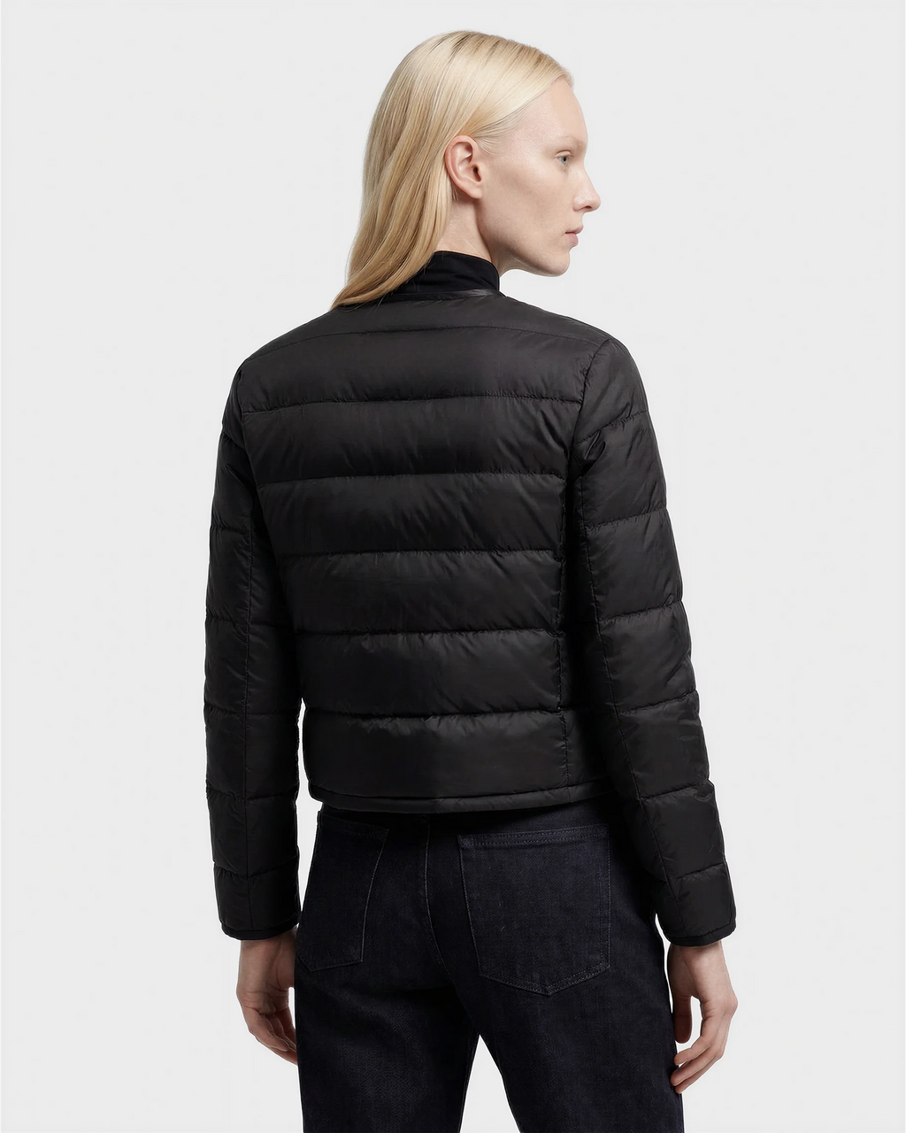 A person with long blond hair, seen from the back, wears the MARKLE - 8125033 Black women’s cropped down jacket by Rudsak with vegan leather trim and dark jeans against a plain light background.