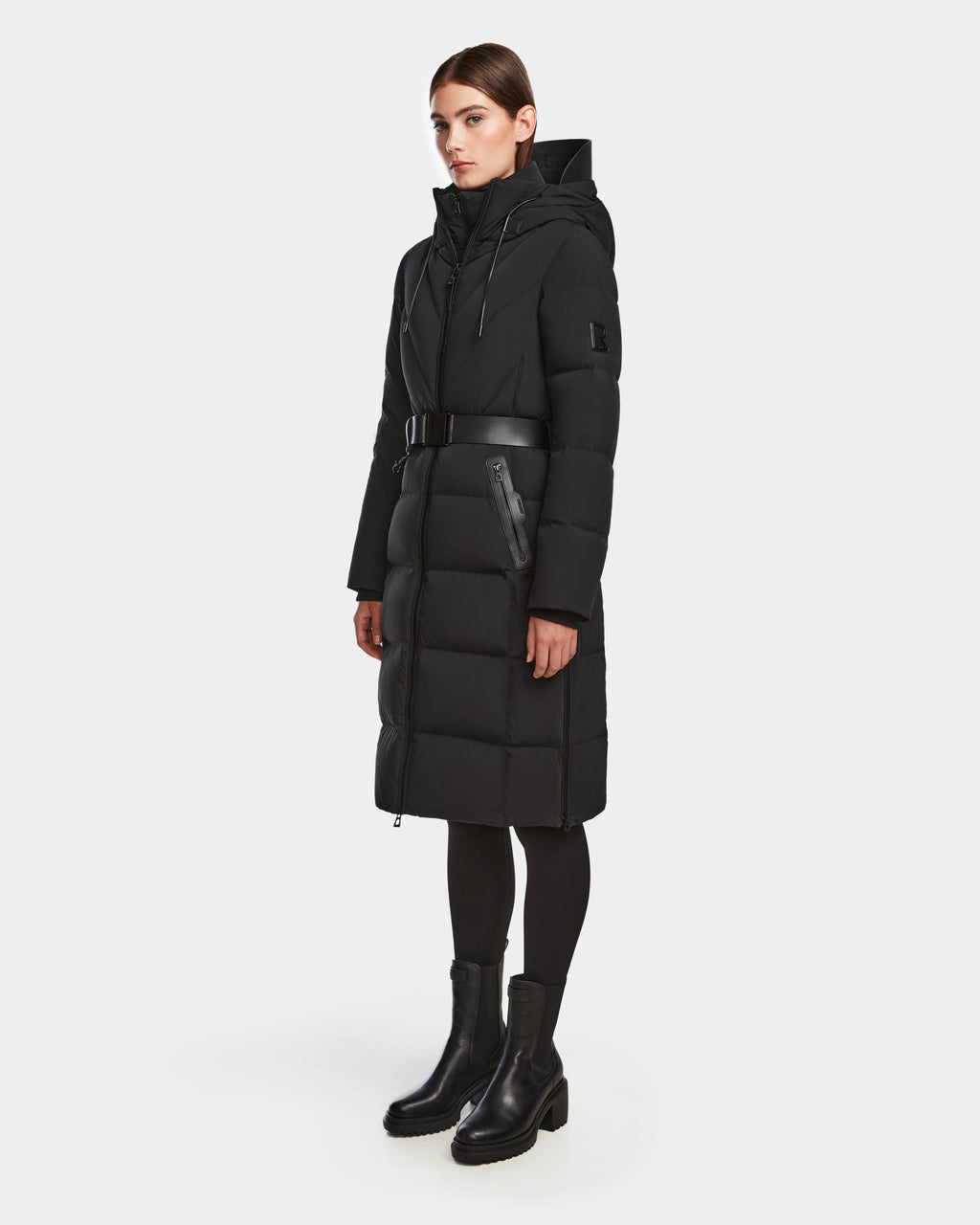 A woman wears the Rudsak DALSA - 8124691 Black long down puffer winter coat with a hood and belted waist, paired with black leggings and ankle boots, against a plain white background.