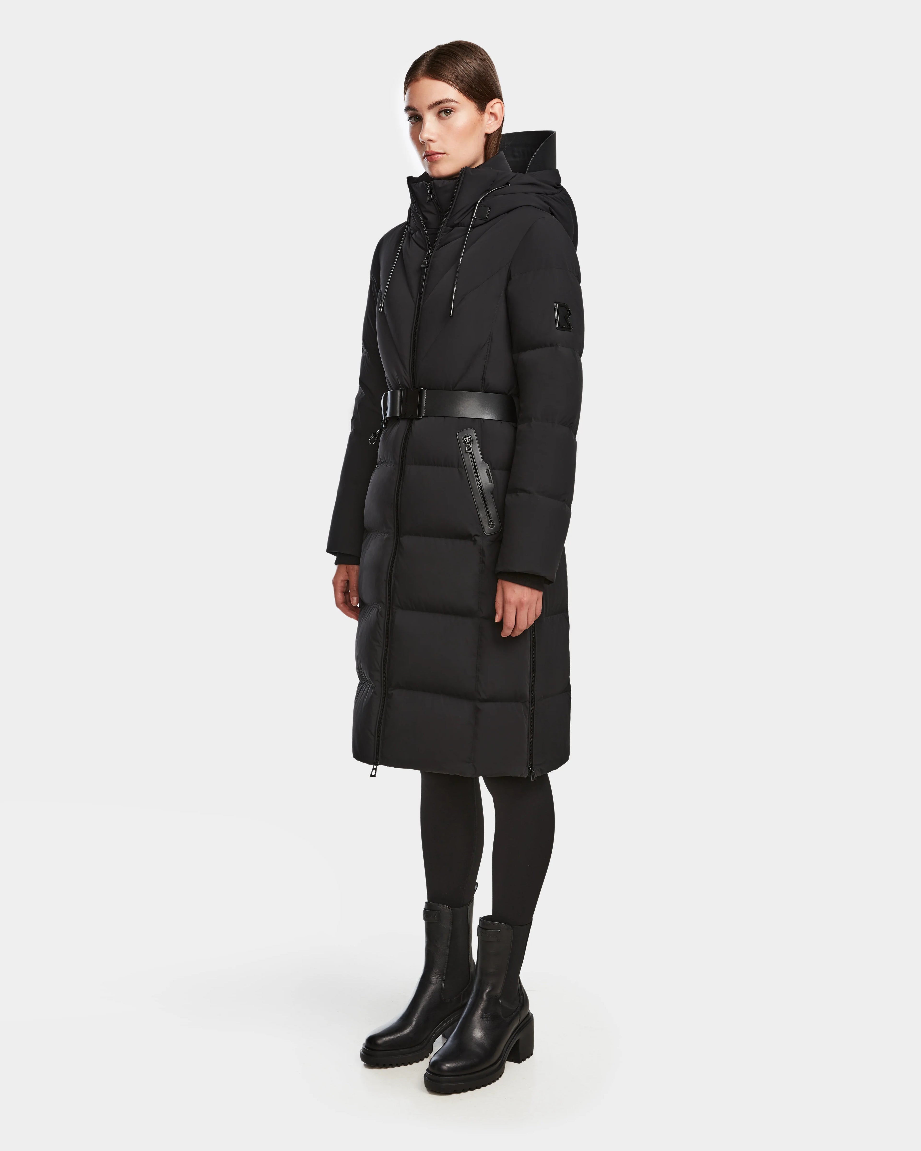 A woman wears the Rudsak DALSA - 8124691 Black long down puffer winter coat with a hood and belted waist, paired with black leggings and ankle boots, against a plain white background.