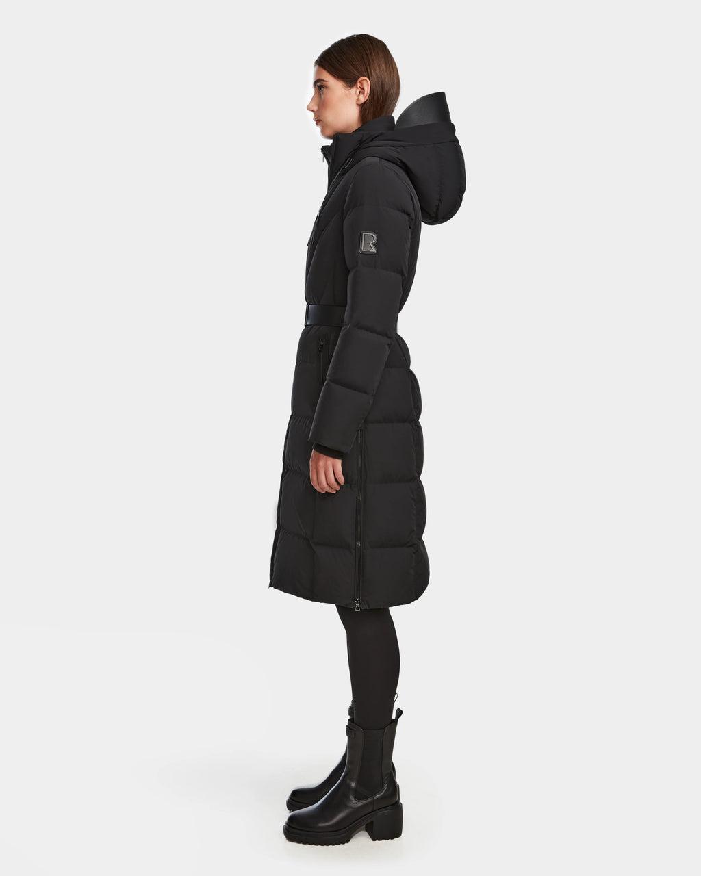 A woman stands in profile wearing the Rudsak DALSA - 8124691 Black long-length down puffer with a hood, black leggings, and black heeled boots against a plain white background.