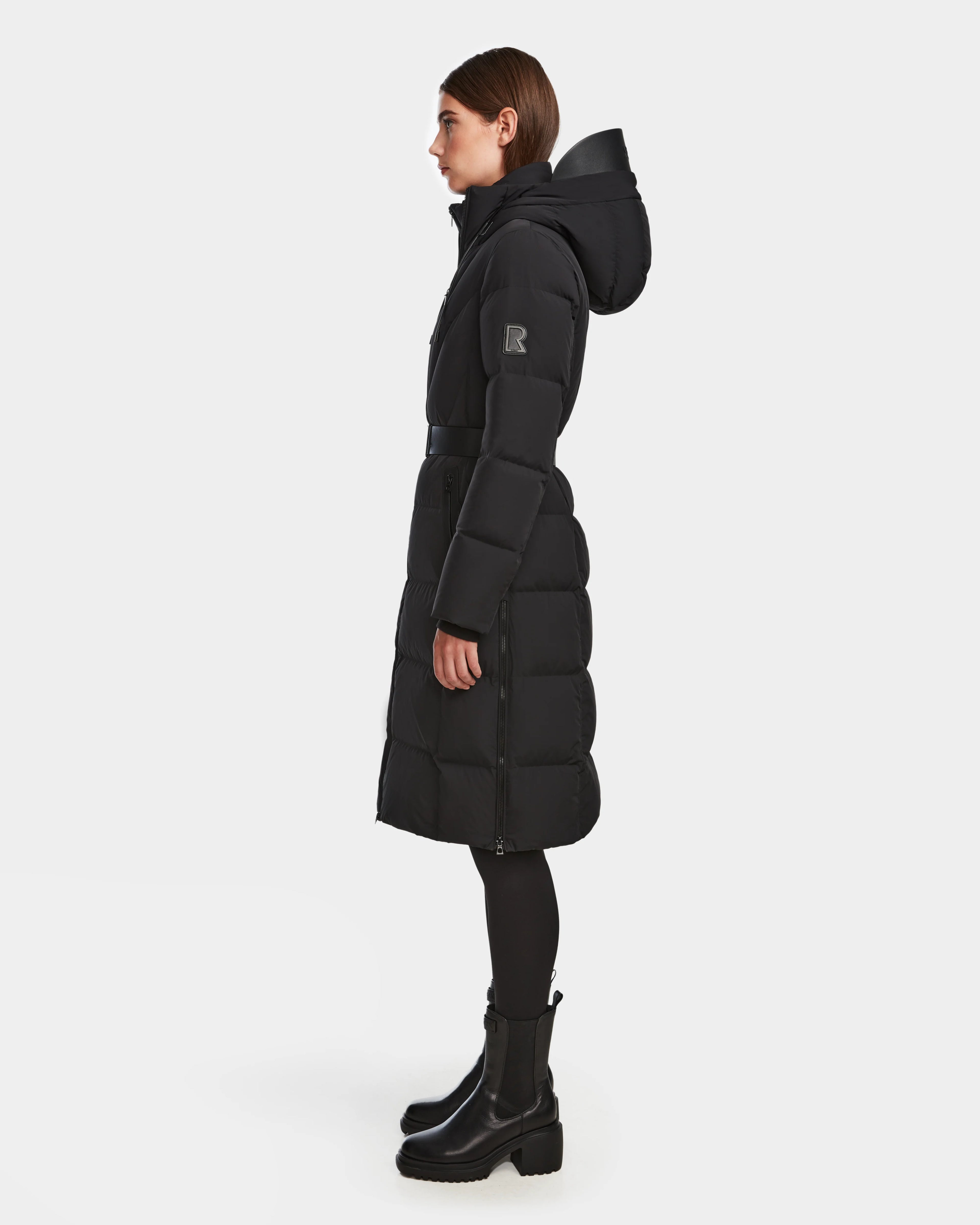 A woman stands in profile wearing the Rudsak DALSA - 8124691 Black long-length down puffer with a hood, black leggings, and black heeled boots against a plain white background.