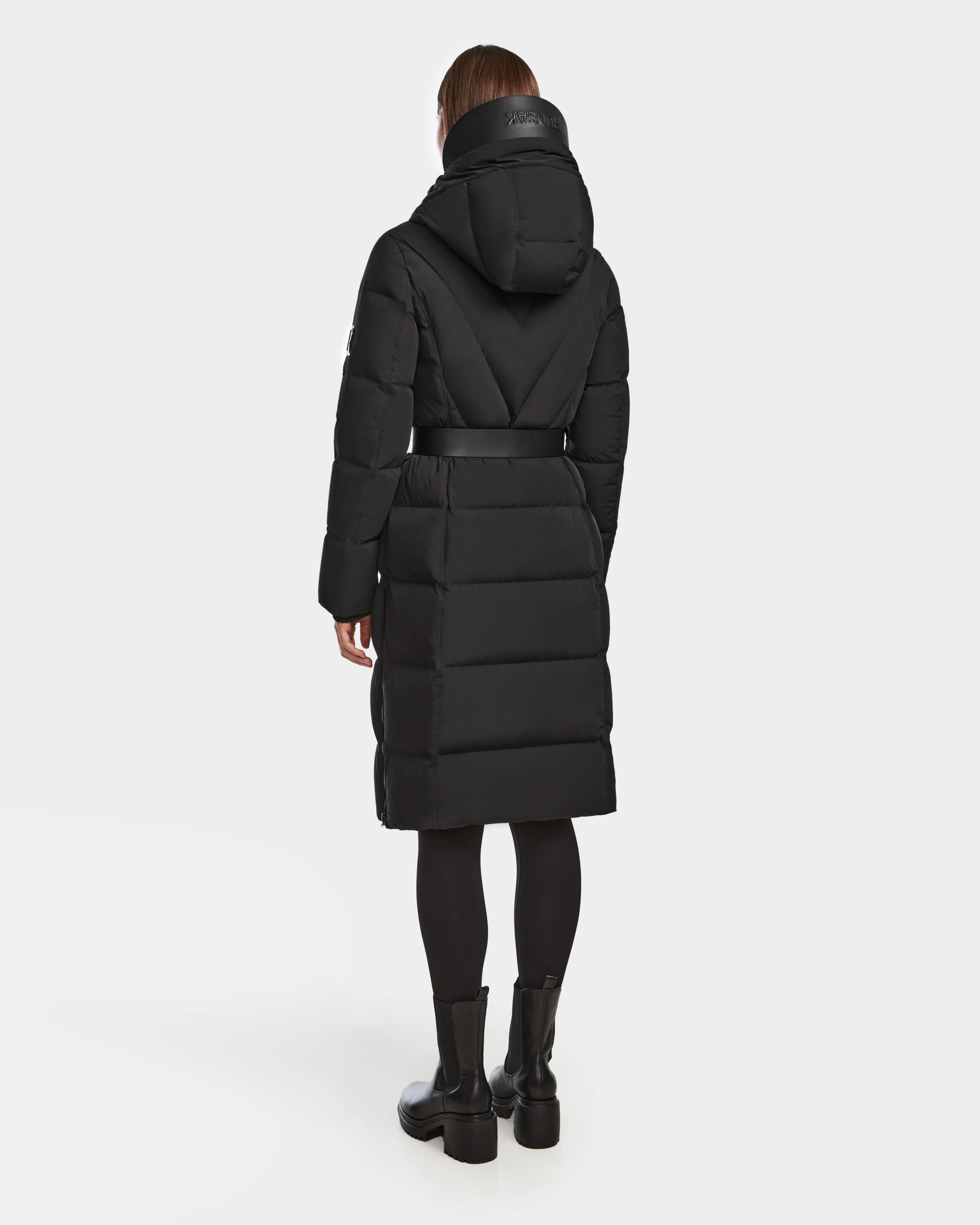 A person is shown wearing the Rudsak DALSA - 8124691 Black women’s long-length down puffer with a hood and belt, paired with black leggings and boots, standing and facing away against a plain white background.