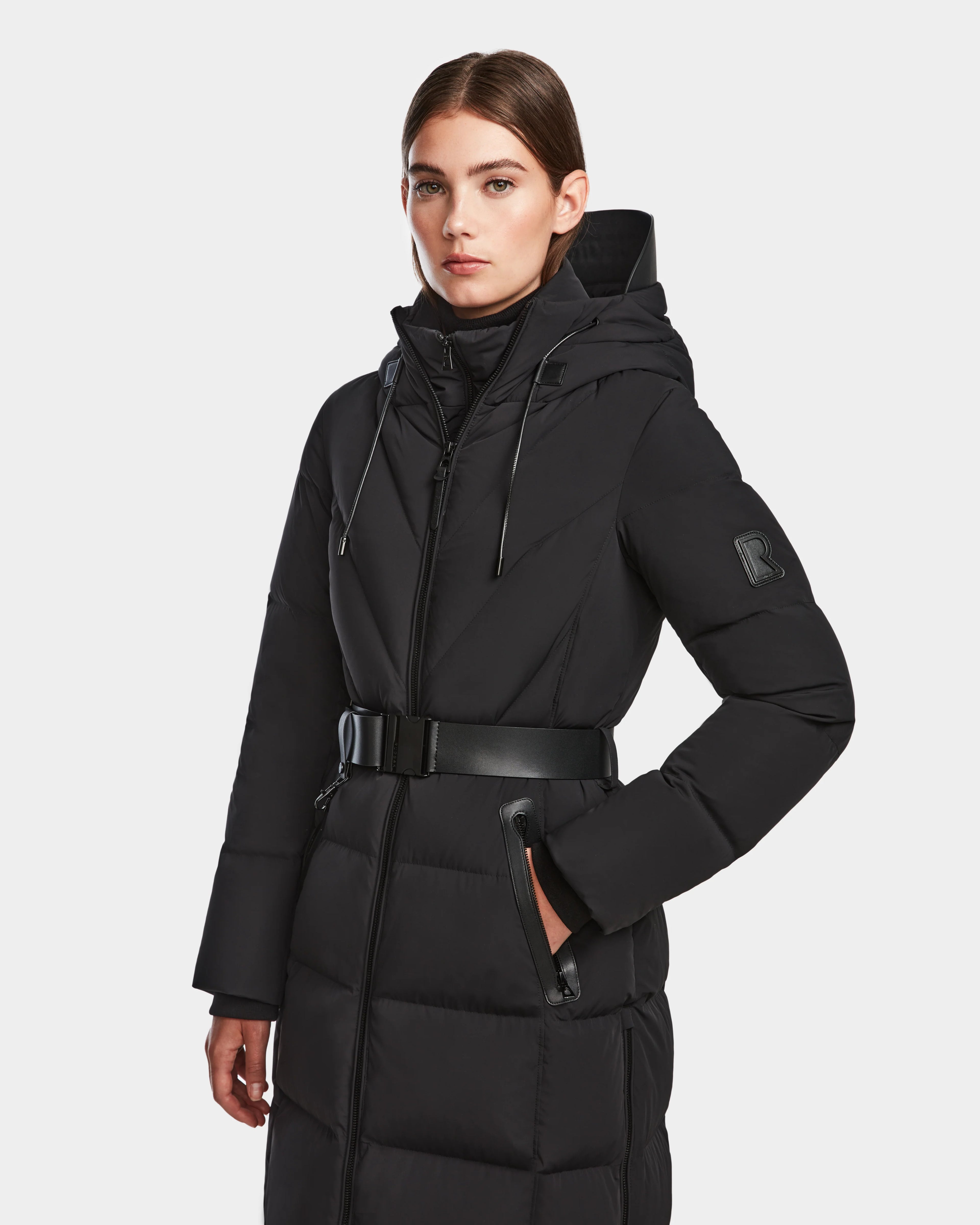 A woman stands with one hand in her pocket, wearing the Rudsak DALSA - 8124691 Black womens long-length down puffer winter coat with a hood, photographed against a plain white background.