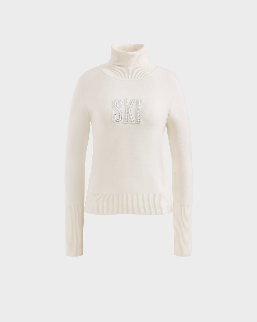 The Rudsak NOVIE - 8525659 Vanilla is a women’s white knit turtleneck ski sweater with ribbed cuffs and hem, featuring bold, textured SKI lettering on the front, shown against a plain light background.