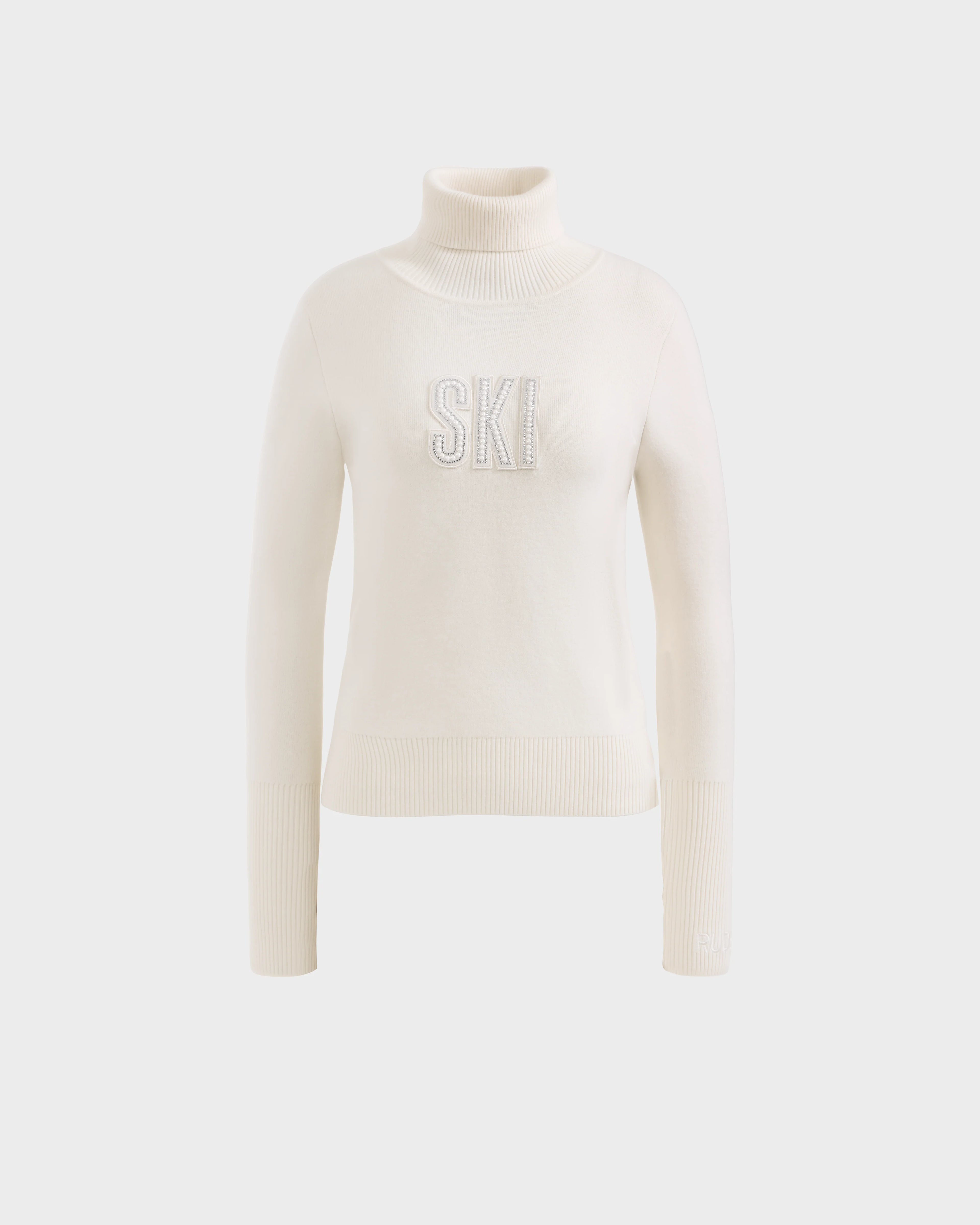 The Rudsak NOVIE - 8525659 Vanilla is a women’s white knit turtleneck ski sweater with ribbed cuffs and hem, featuring bold, textured SKI lettering on the front, shown against a plain light background.
