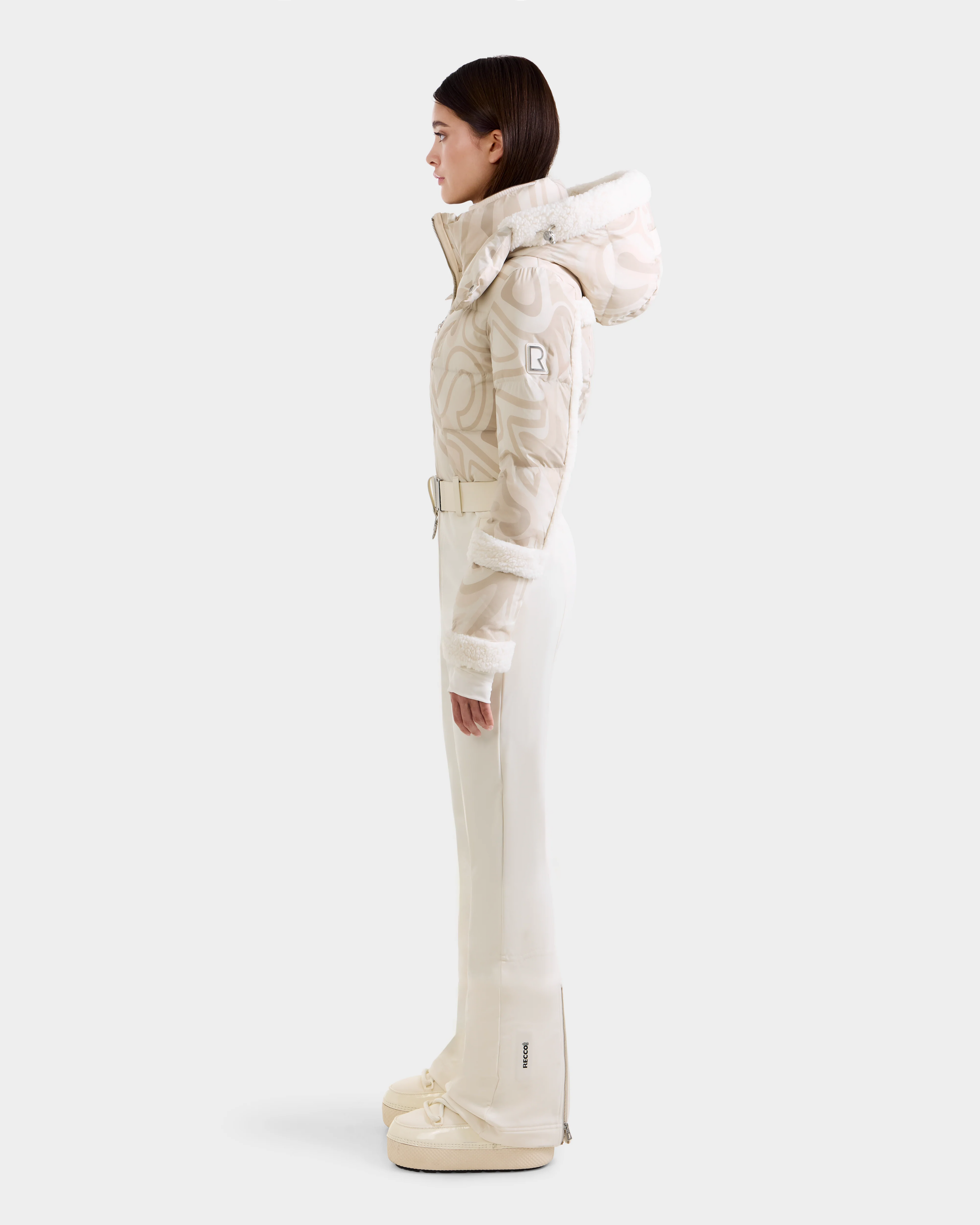 A woman in profile wears the Rudsak CARMEN P - 8125707 Vanilla Deja vu ski suit—featuring a high collar, fur trim, belted waist, flared pants, and matching boots—available for pre-order and shipping by November 30th.
