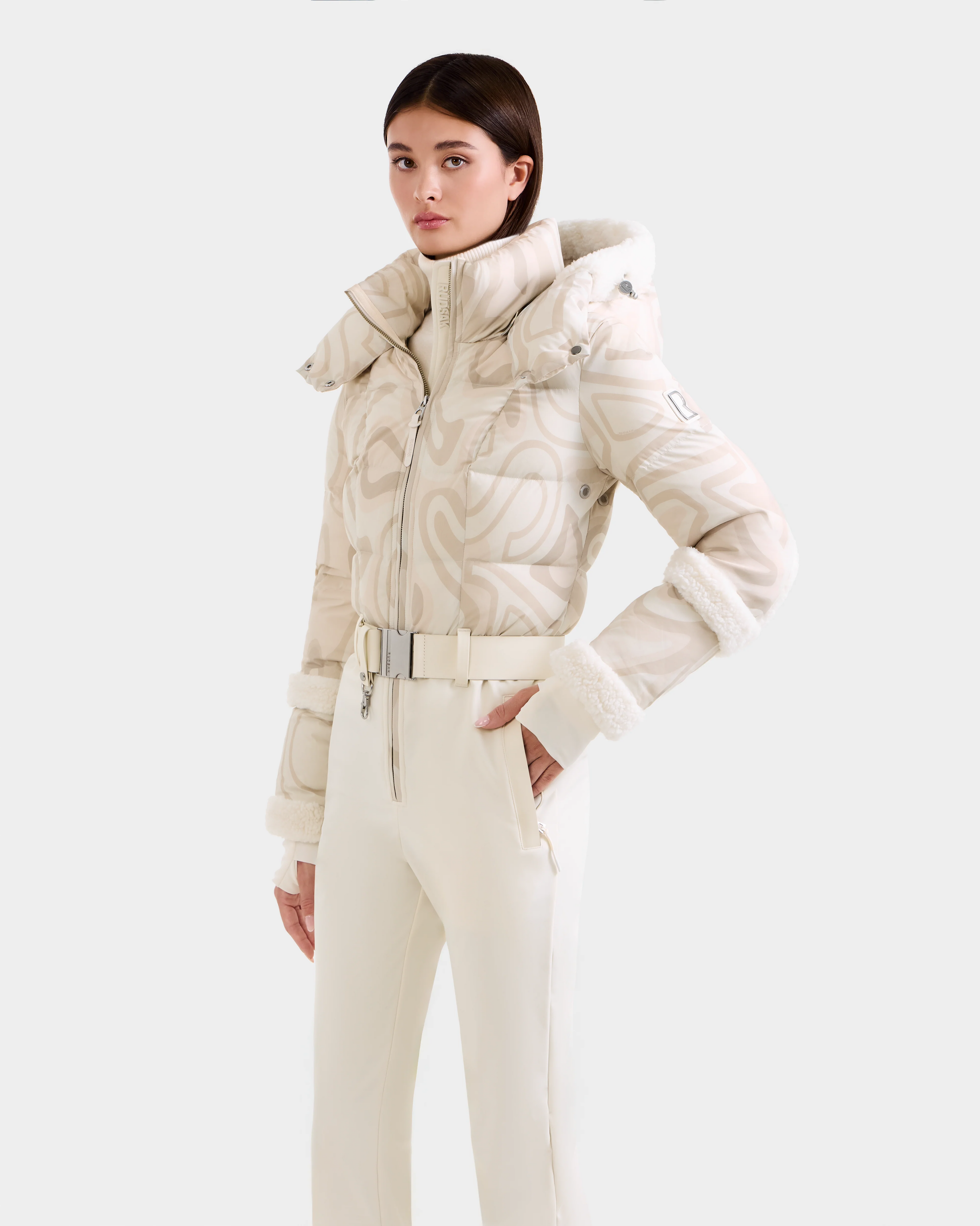 A woman models the Rudsak CARMEN P - 8125707 Vanilla Deja vu off-white patterned puffer jacket with fur trim and matching high-waisted pants, standing hands-in-pockets against a light background.