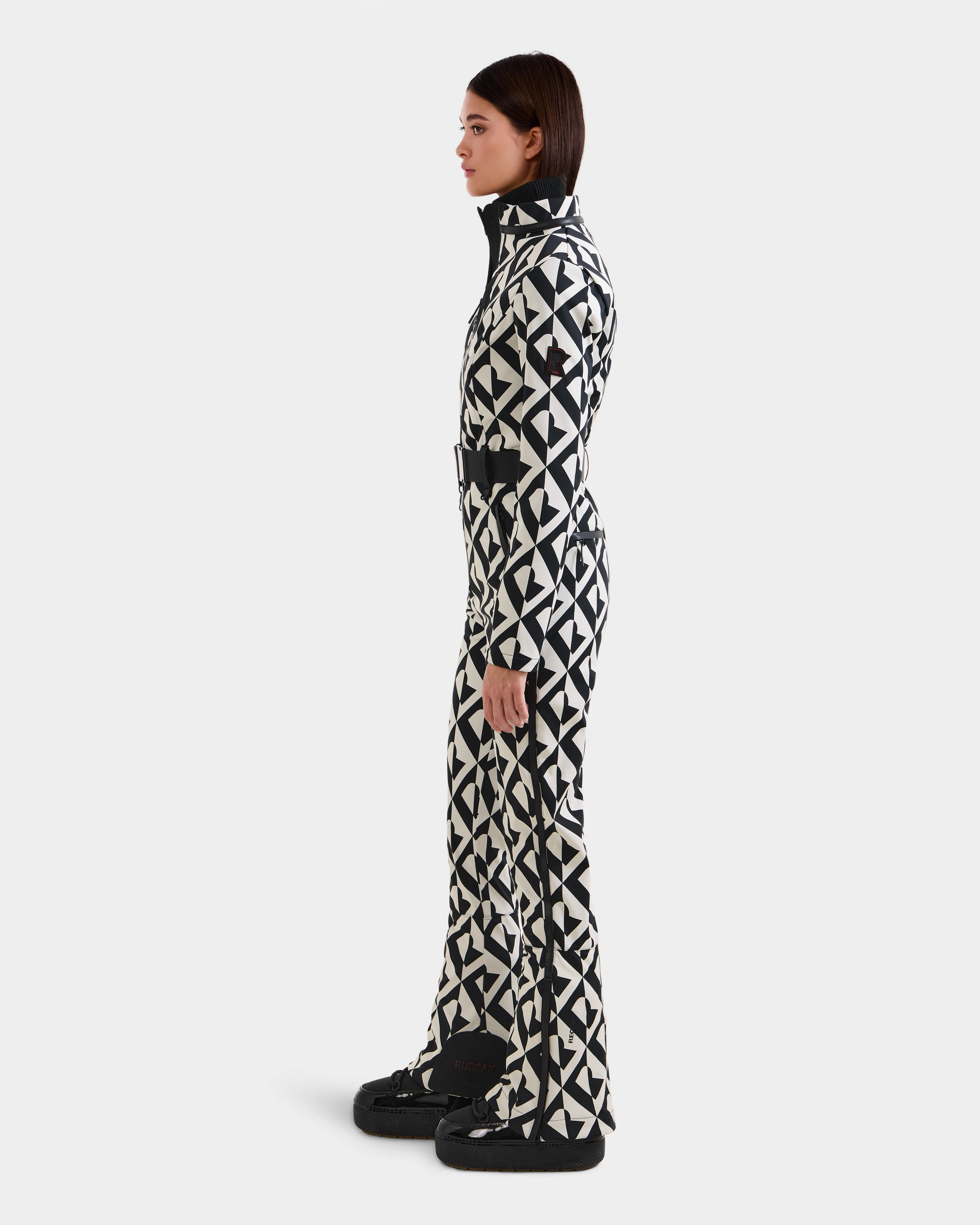 A woman stands in profile against a white background, wearing the Rudsak JOSIE - 8124817 Vanilla Monogram women's ski suit with a high collar and belt, paired with black snow boots.