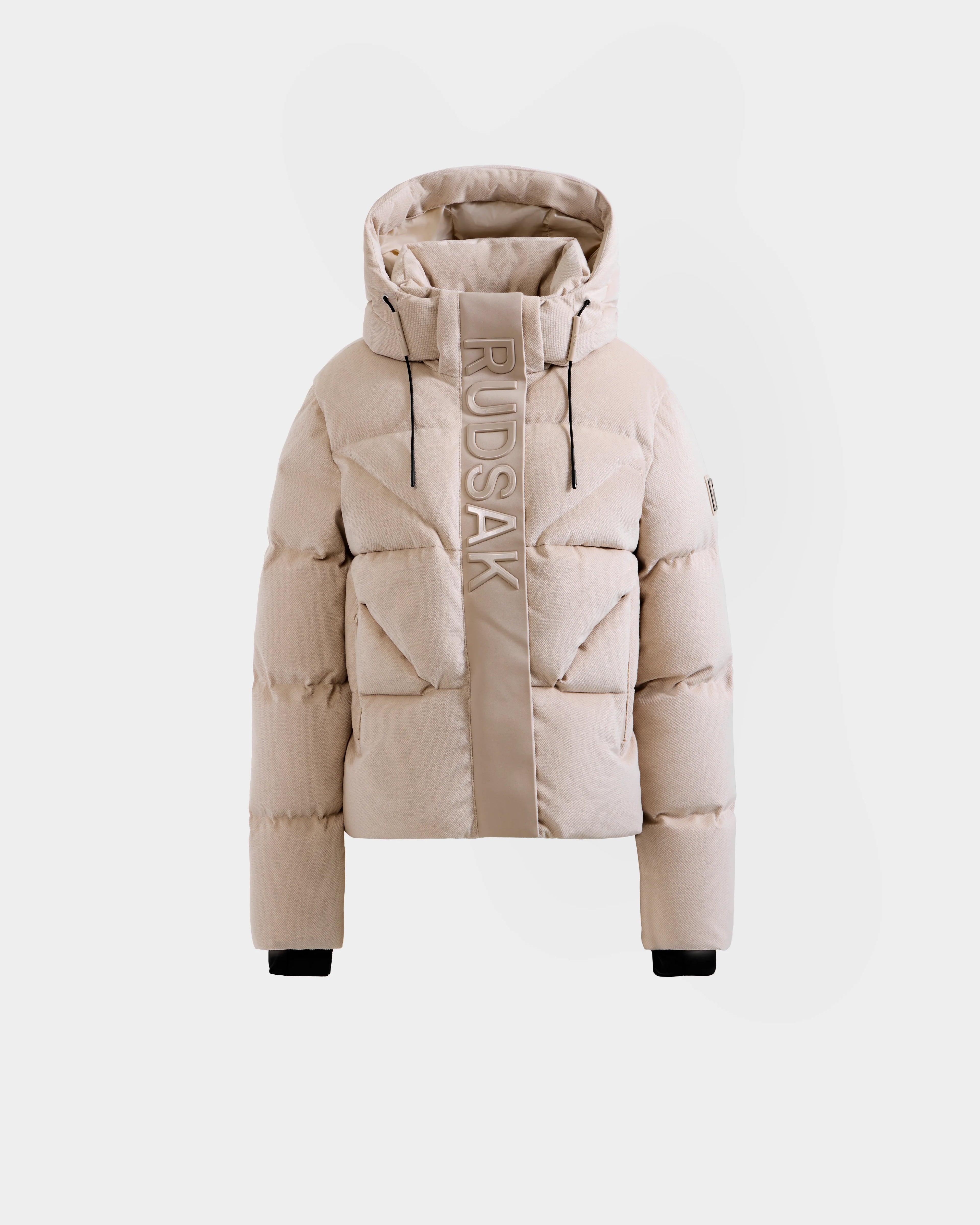 The STEFANIE V - L125584 Vanilla by Rudsak is a beige down puffer jacket with a hood, black wrist cuffs, vertical Rudsak branding on the left front, front zipper closure, and a modern quilted design.