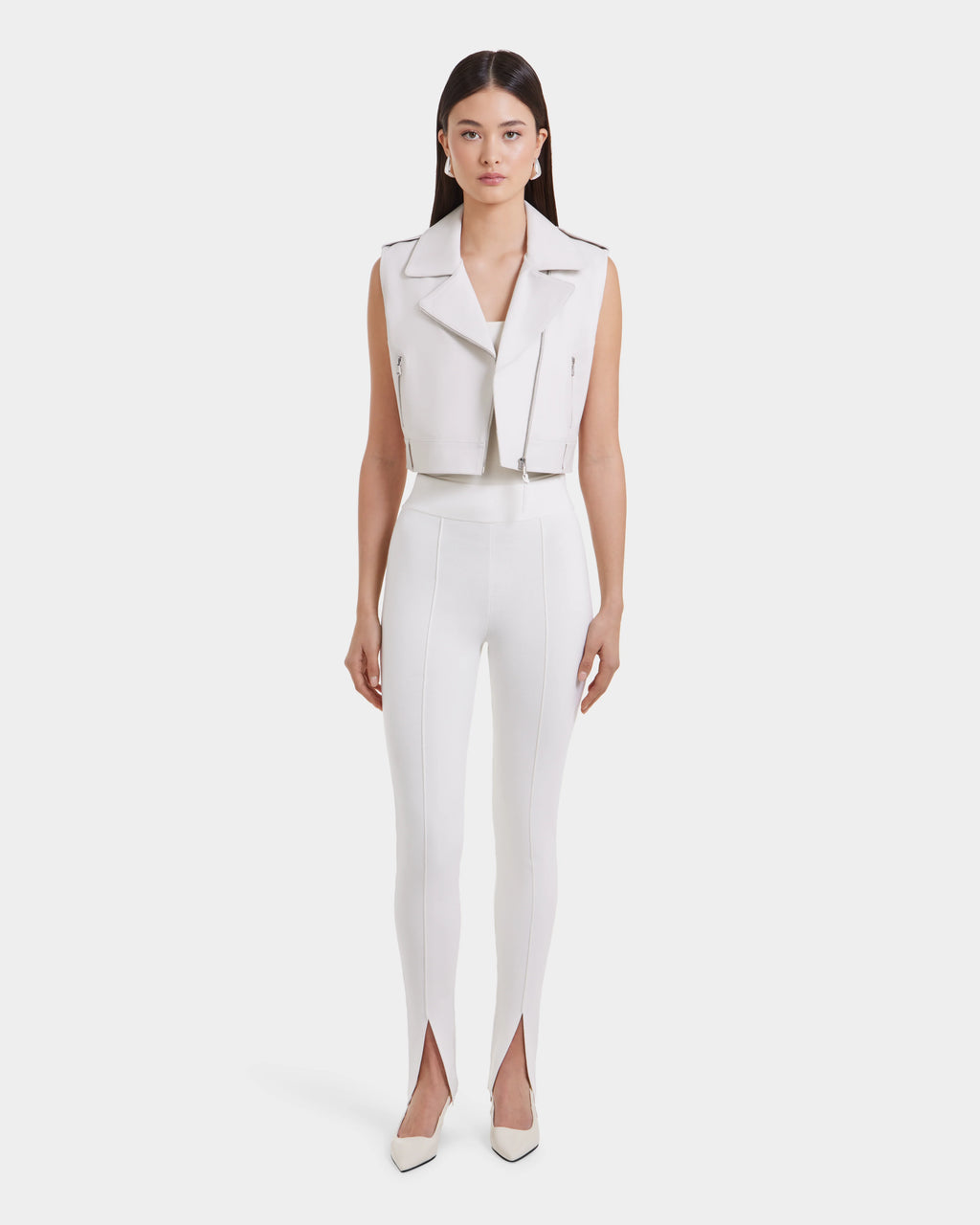 A woman stands facing forward in the Rudsak BETTY - 8125000 Optic White sleeveless leather vest, high-waisted white pants with ankle front slits, and white pointed-toe shoes against a plain white background.