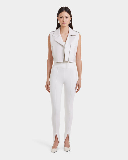 A woman stands facing forward in the Rudsak BETTY - 8125000 Optic White sleeveless leather vest, high-waisted white pants with ankle front slits, and white pointed-toe shoes against a plain white background.
