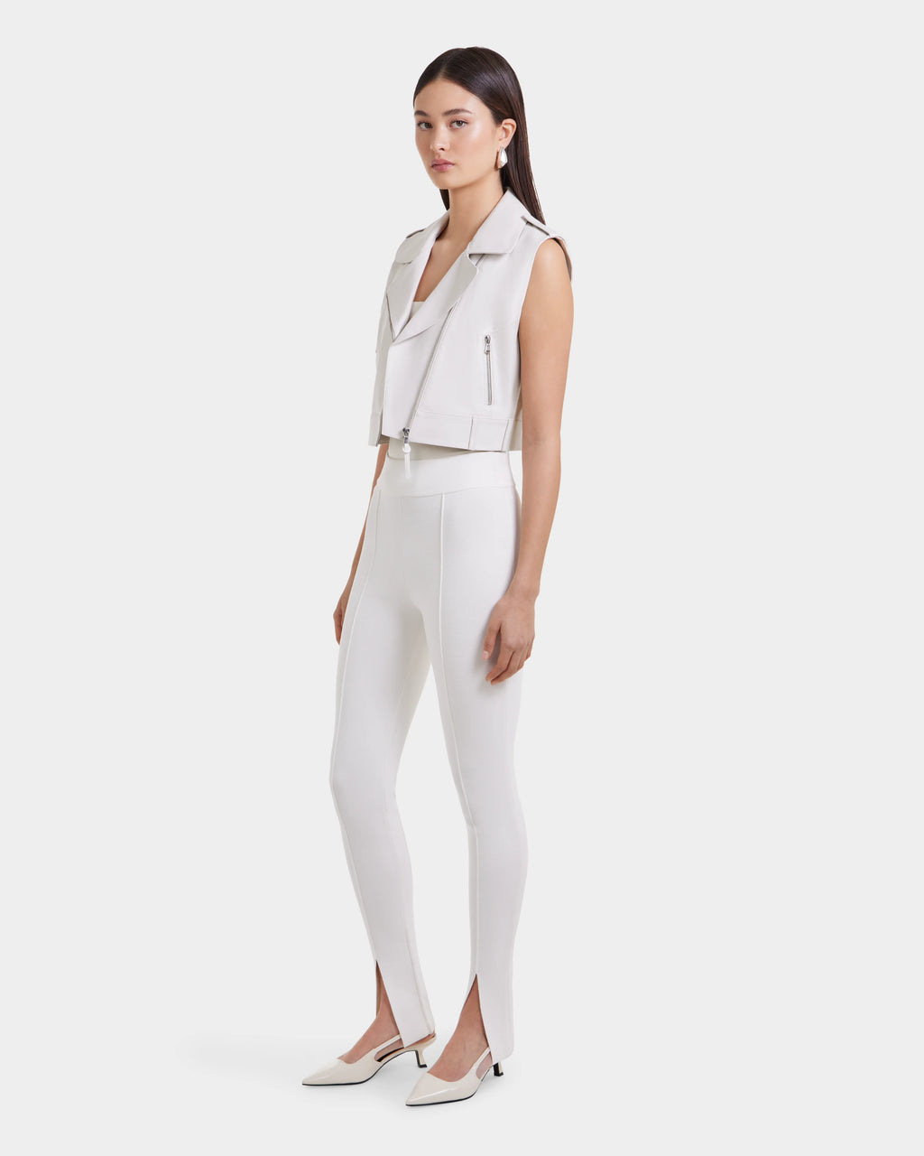 A woman with straight dark hair and a neutral expression wears the BETTY - 8125000 Optic White sleeveless moto vest by Rudsak, paired with high-waisted white pants featuring front ankle slits and white pointed-toe heels against a plain background.