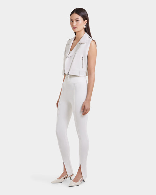 A woman with straight dark hair and a neutral expression wears the BETTY - 8125000 Optic White sleeveless moto vest by Rudsak, paired with high-waisted white pants featuring front ankle slits and white pointed-toe heels against a plain background.
