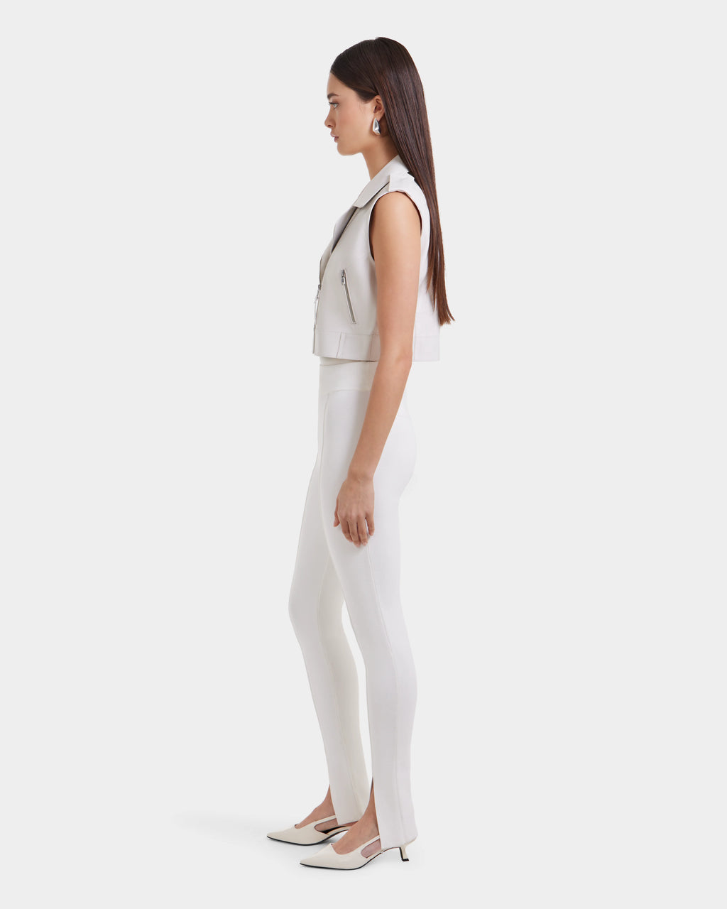 A woman stands in profile against a white background, wearing the BETTY - 8125000 Optic White sleeveless cropped leather vest by Rudsak, paired with fitted white pants and pointed-toe heels. Her straight brown hair is worn down.