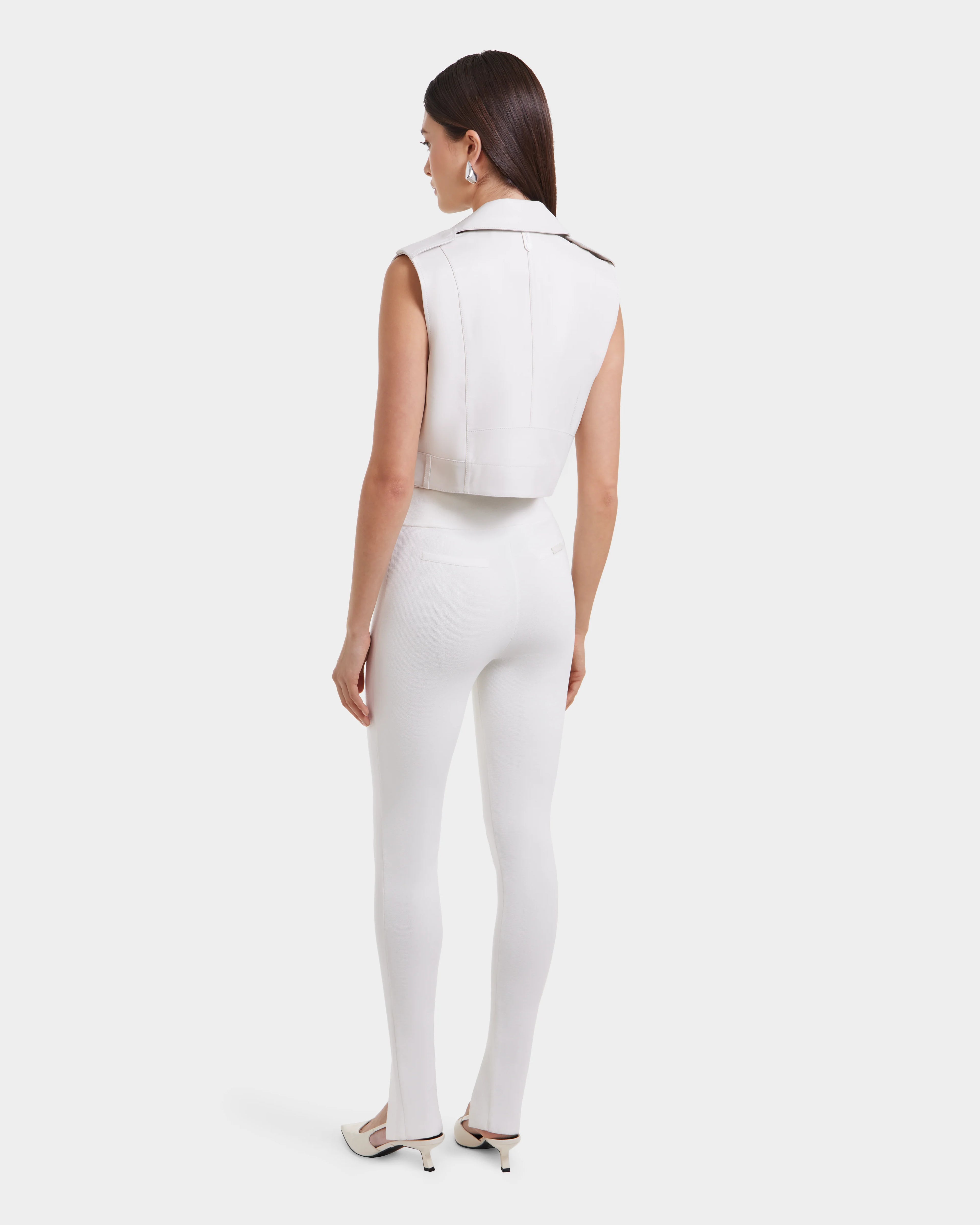 A woman with straight dark hair and hoop earrings faces away, wearing the Rudsak BETTY - 8125000 Optic White cropped leather vest and fitted white pants with heeled shoes, set against a plain light background.