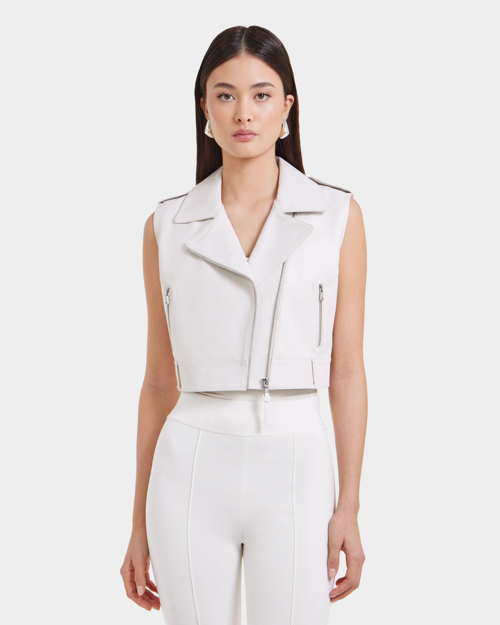 A woman with straight dark hair models the Rudsak BETTY - 8125000 Optic White high-waisted pants and a cropped moto leather vest, standing against a light background for a modern, cool vibe.